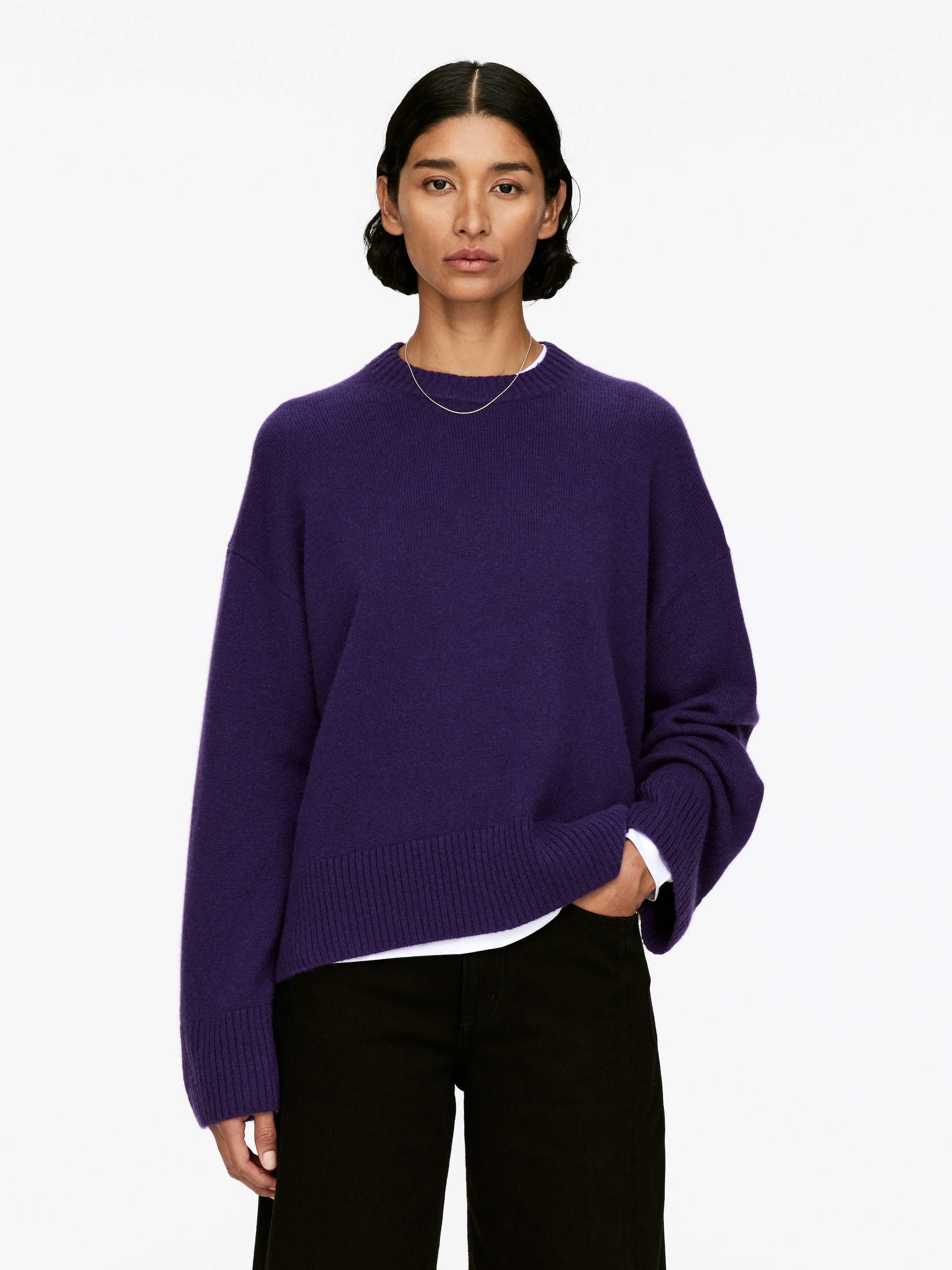 Ingrandisci l'immagine: Un maglione oversize in puro cashmere di colore viola scuro, con scollo rotondo e polsini e orlo a coste, è indossato dalla modella. Una camicia bianca a maniche lunghe è stratificata sotto, visibile all'orlo e ai polsini, e il maglione è abbinato a pantaloni neri.