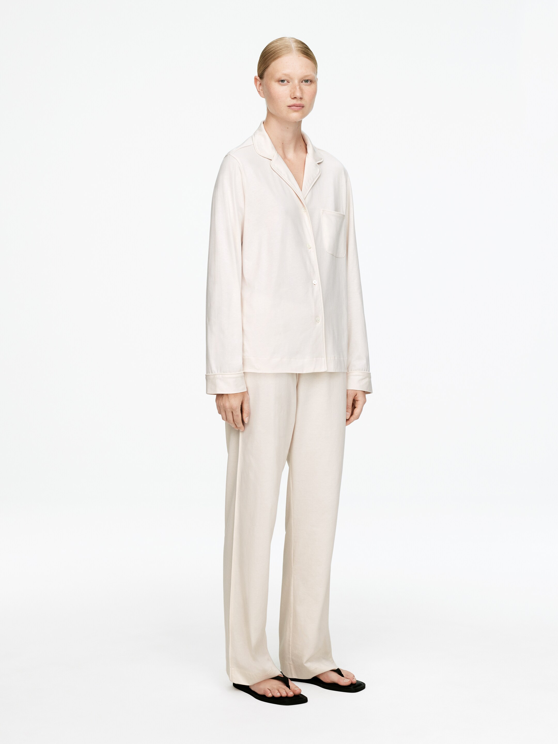 Agrandir l'image: Un ensemble de pyjama beige clair est présenté de face avec un léger angle. Le haut à manches longues présente une fermeture à boutons avec des revers crantés et une poche plaquée unique. Le pantalon assorti offre une coupe droite et décontractée.