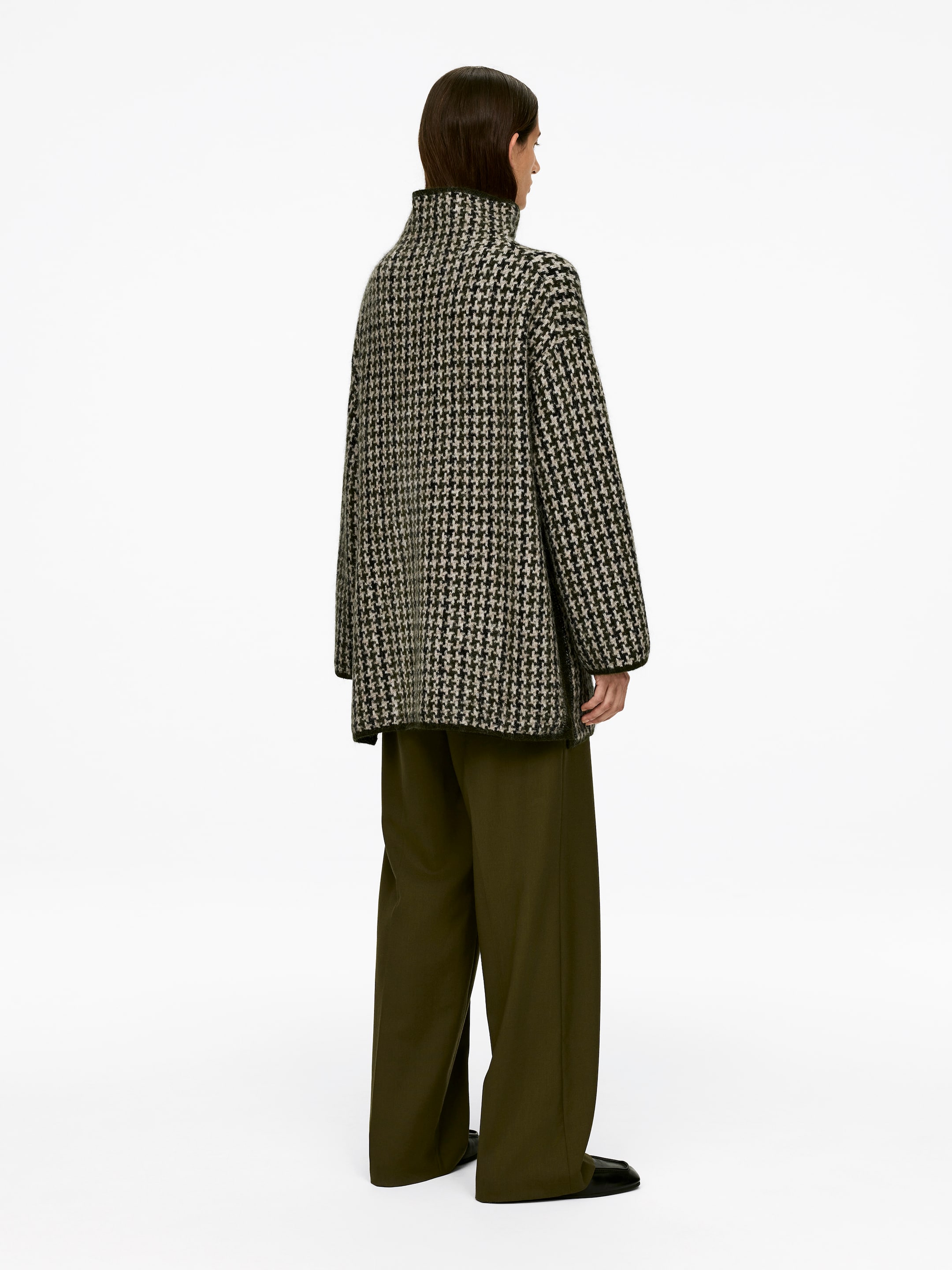Visa större bild: En långärmad, avslappnad passform jacquardstickad tröja med ett svart och offwhite hundtandsmönster och en mock hals, bärs av modellen. Tröjan är stylad med mörkgröna byxor med breda ben.