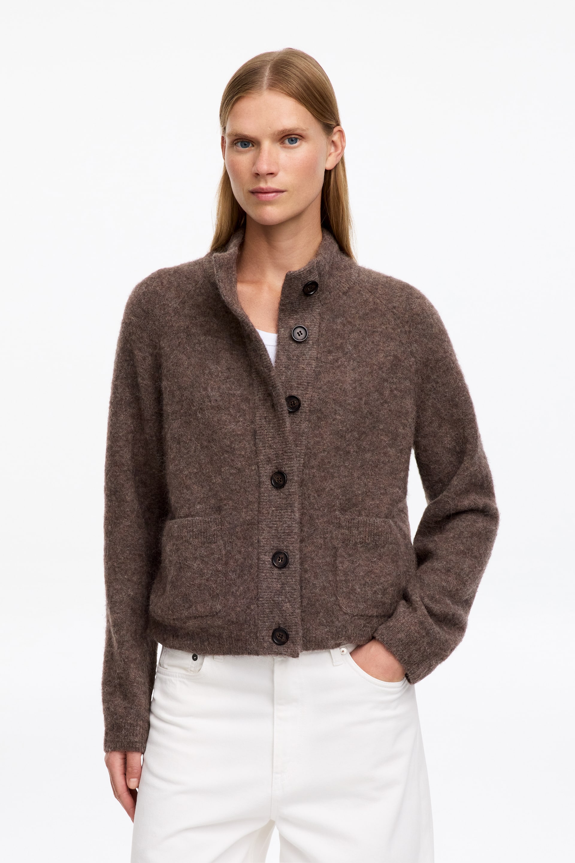 Größeres Bild ansehen: Eine braune, strukturierte Strickjacke wird vom Model getragen und mit einer weißen Hose sowie einem weißen T-Shirt kombiniert. Die Strickjacke verfügt über einen hohen Kragen, eine durchgehende Knopfleiste und aufgesetzte Fronttaschen.