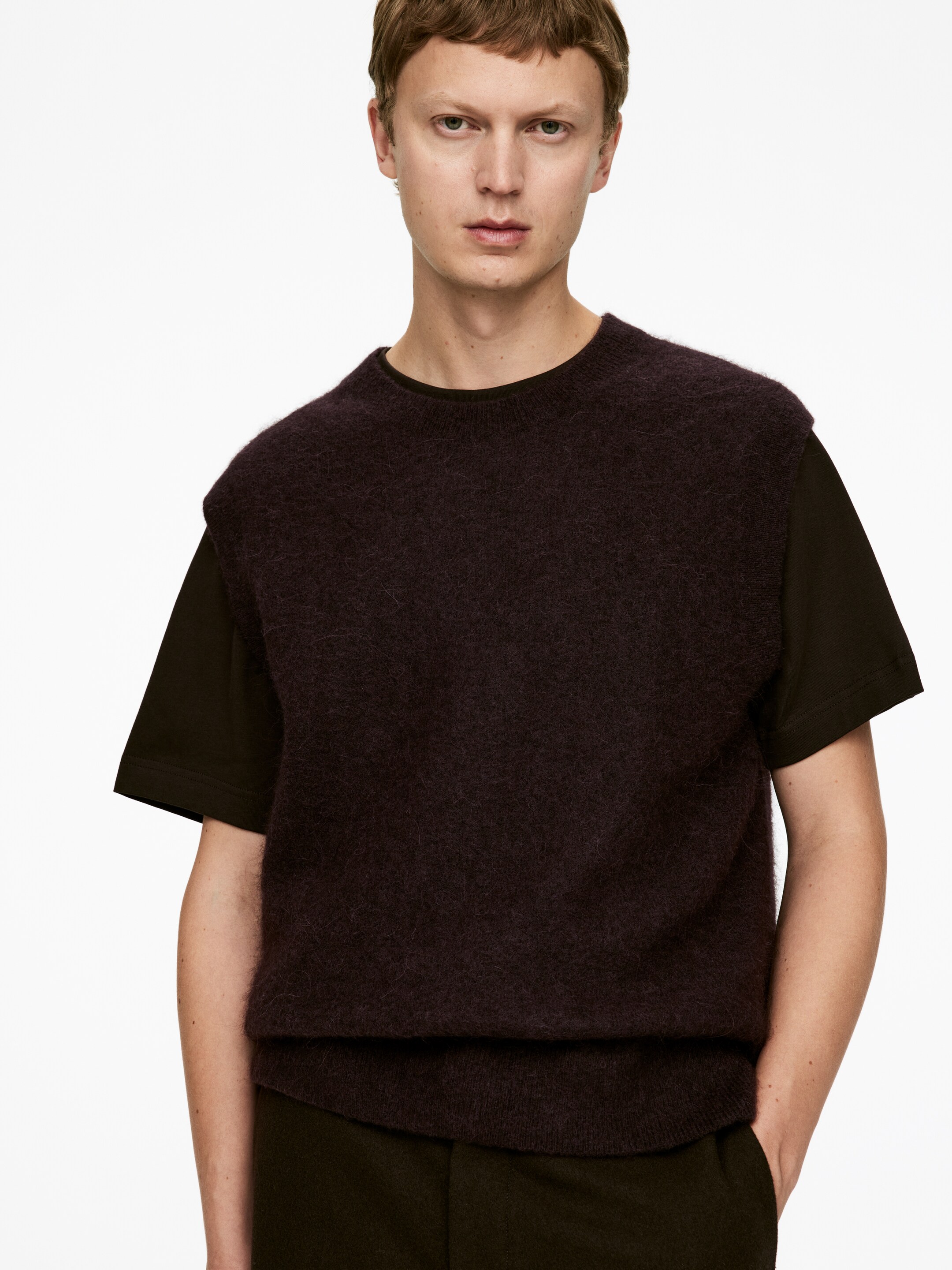 Grotere afbeelding bekijken: Een donker pruimkleurige, met een wazige textuur mouwloze gebreide vest wordt gedragen, naar voren gericht, over een donker t-shirt met korte mouwen. De vest heeft een ronde hals en een geribde zoom, en biedt een ontspannen pasvorm.