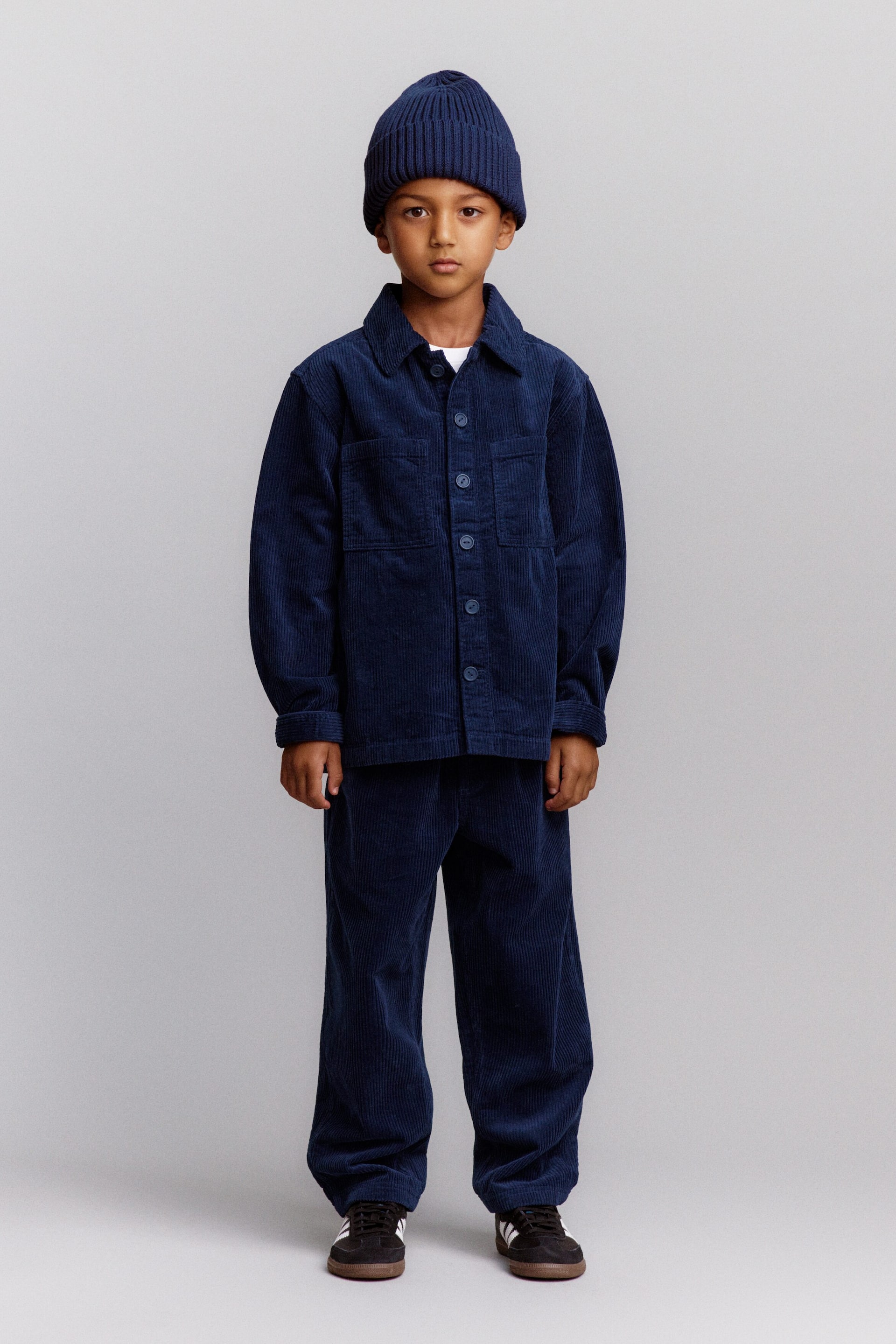 Visualizza immagine più grande: Un bambino indossa un outfit coordinato blu navy composto da un berretto in cotone a coste con risvolto, una overshirt in corduroy a coste larghe dal fit ampio e pantaloni dal taglio rilassato. Inquadrata frontalmente, l’overshirt presenta colletto classico, chiusura con bottoni sul davanti e tasche applicate sul petto, mentre i pantaloni coordinati cadono in una vestibilità rilassata e affusolata.