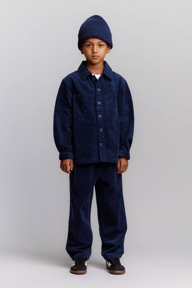 Ein Kind trÃ¤gt ein farblich abgestimmtes marineblaues Outfit mit einer gerippten Baumwoll-MÃ¼tze mit umgeschlagenem Saum, einem weit geschnittenen Cord-Ãberhemd mit breitem Cord-Ripp und einer locker geschnittenen Hose. Von vorne gezeigt, verfÃ¼gt das Ãberhemd Ã¼ber einen klassischen Kragen, eine durchgehende Knopfleiste und aufgesetzte Brusttaschen, wÃ¤hrend die passende Hose in einer entspannten, schmal zulaufenden Passform fÃ¤llt. - 1