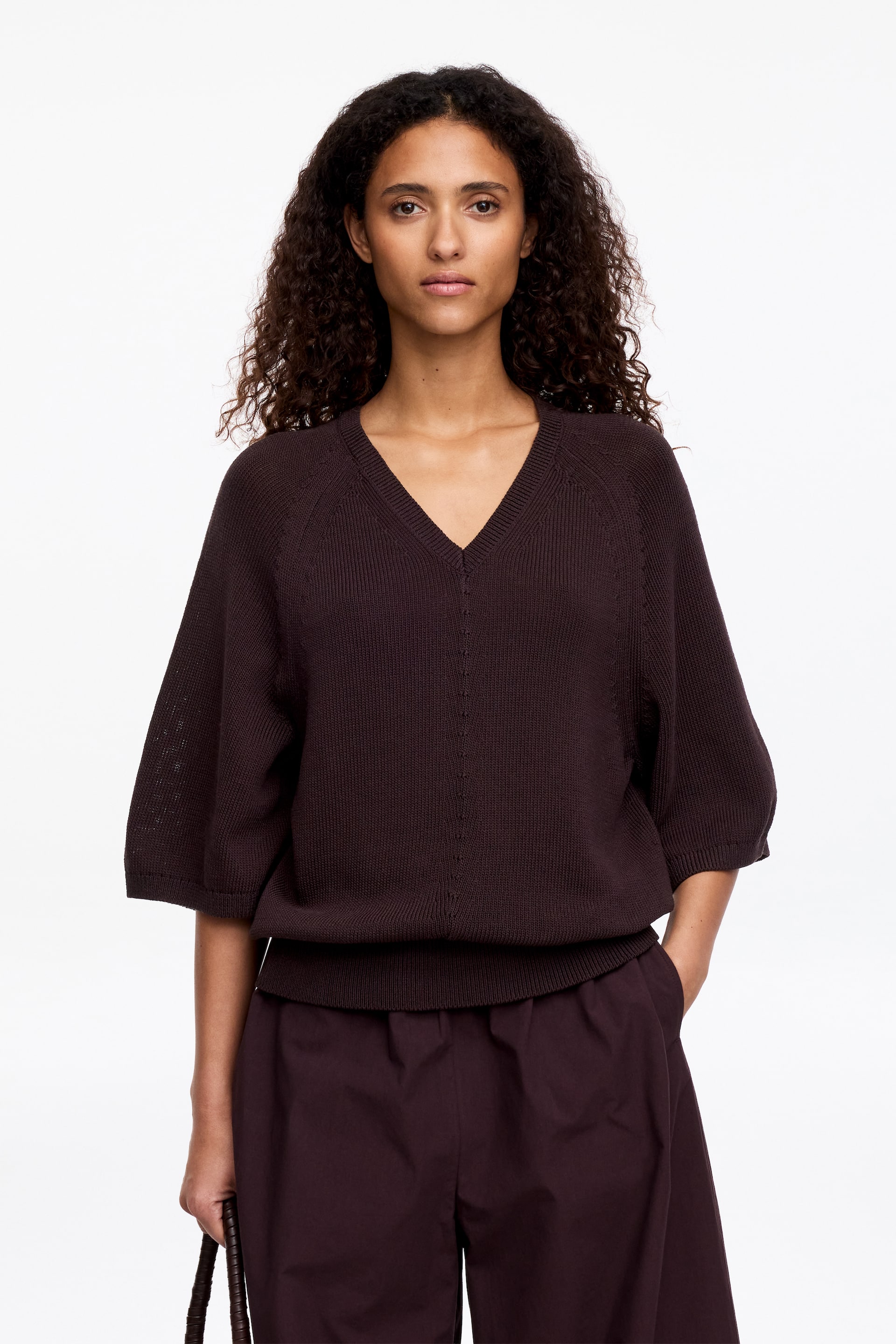 Größeres Bild anzeigen: Ein dunkelbrauner Strickpullover wird von vorne präsentiert, mit einem V-Ausschnitt und weiten, dreiviertellangen Raglanärmeln. Er zeigt einen gerippten V-Ausschnitt, gerippte Ärmelabschlüsse und einen gerippten Saum, mit einem dezenten vertikalen Nahtdetail entlang der Vorderseite.