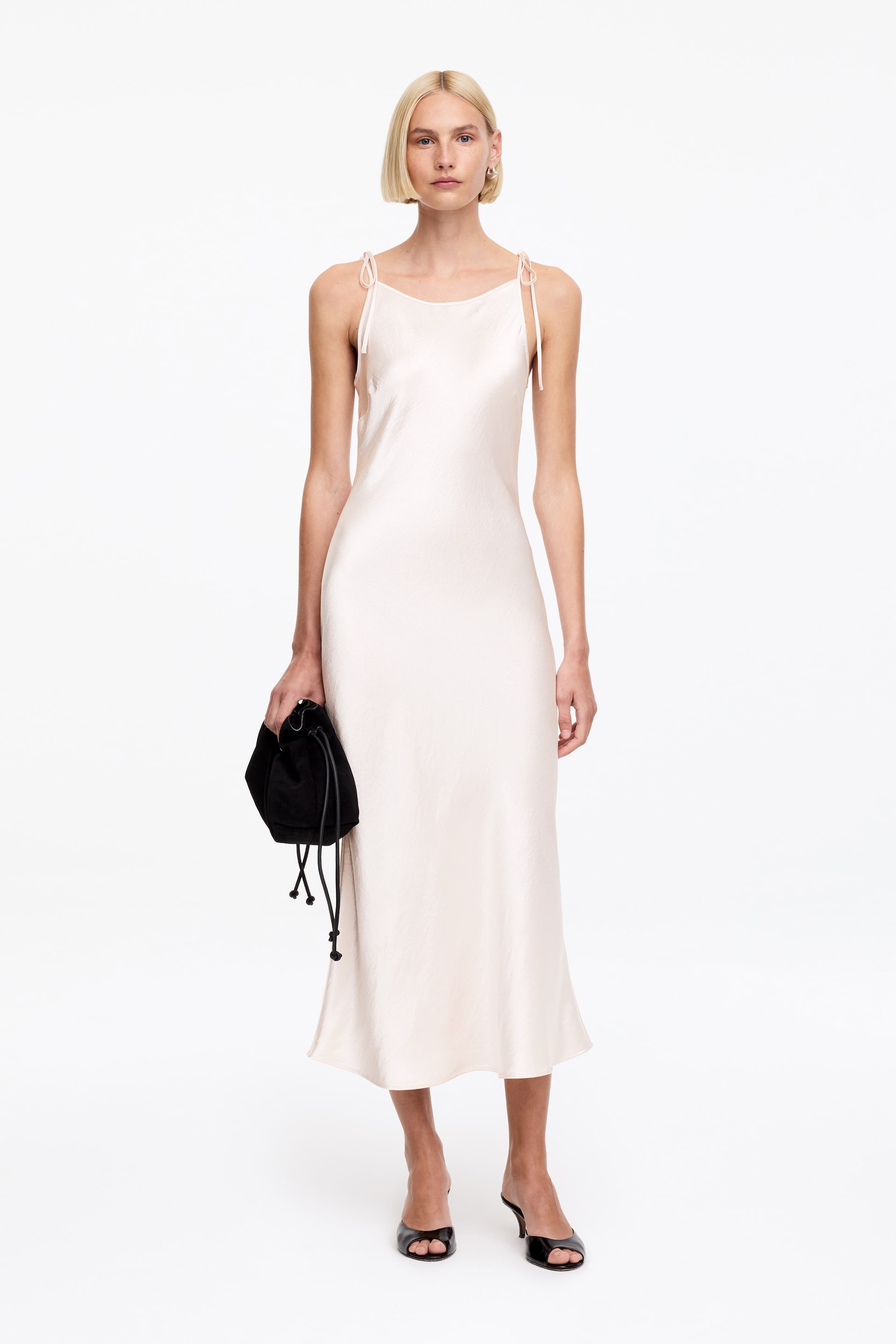 Nagyobb kép megtekintése: Egy világos krémszínű kombiné ruha, amely kerek nyakkivágással és vékony spagettipántokkal rendelkezik, amelyek a vállaknál megkötöttek. A midi hosszúságú ruha selymes anyagból készült, finoman követi a test vonalát, és a vádli közepéig ér.