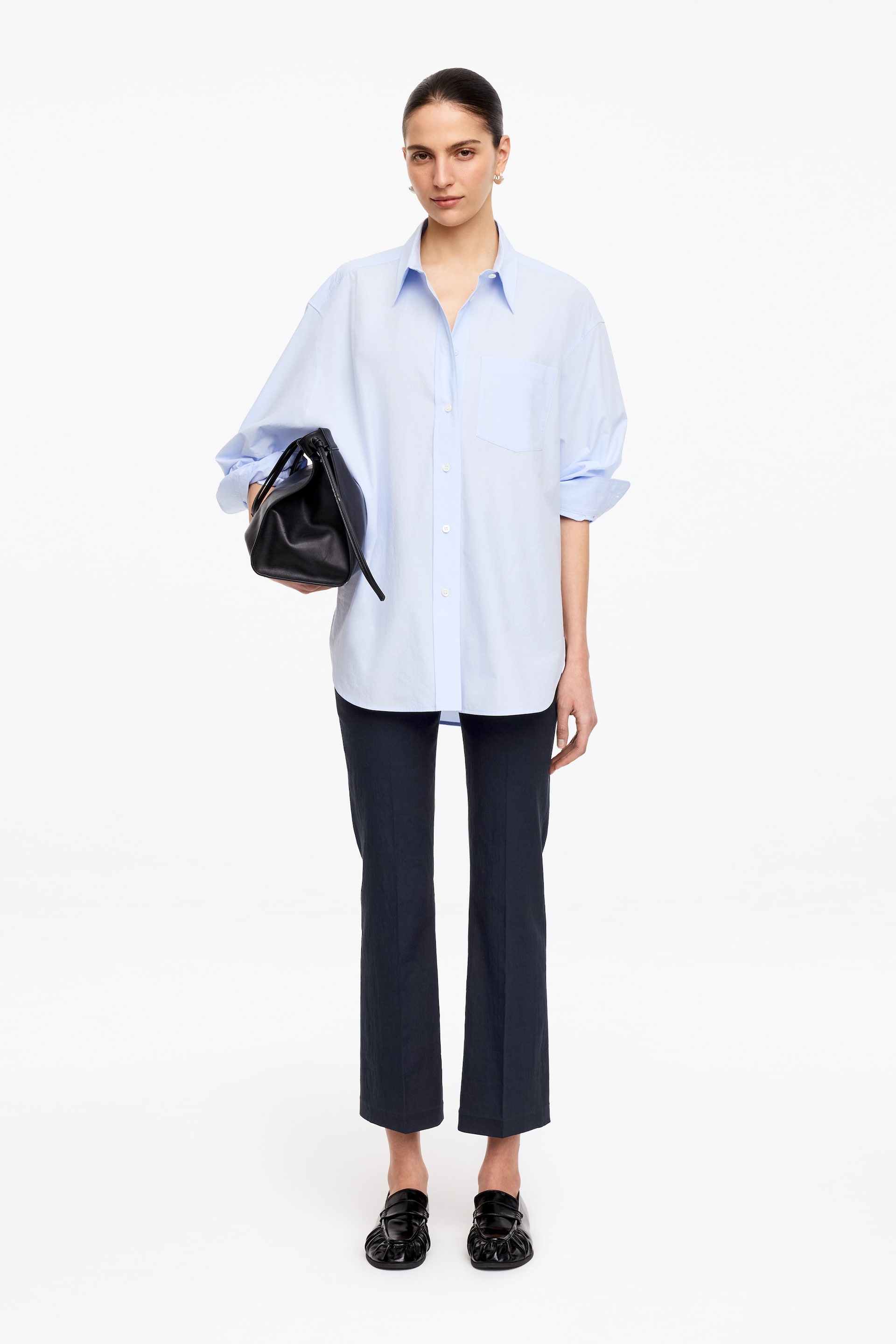 Se større billede: En lyseblå oversized skjorte med knapper har en spids krave, hvide knapper ned langs fronten og en enkelt brystlomme på venstre side. De lange ærmer er rullet op til midt på underarmen, og skjorten er vendt fremad, hvilket fremhæver en afslappet pasform.