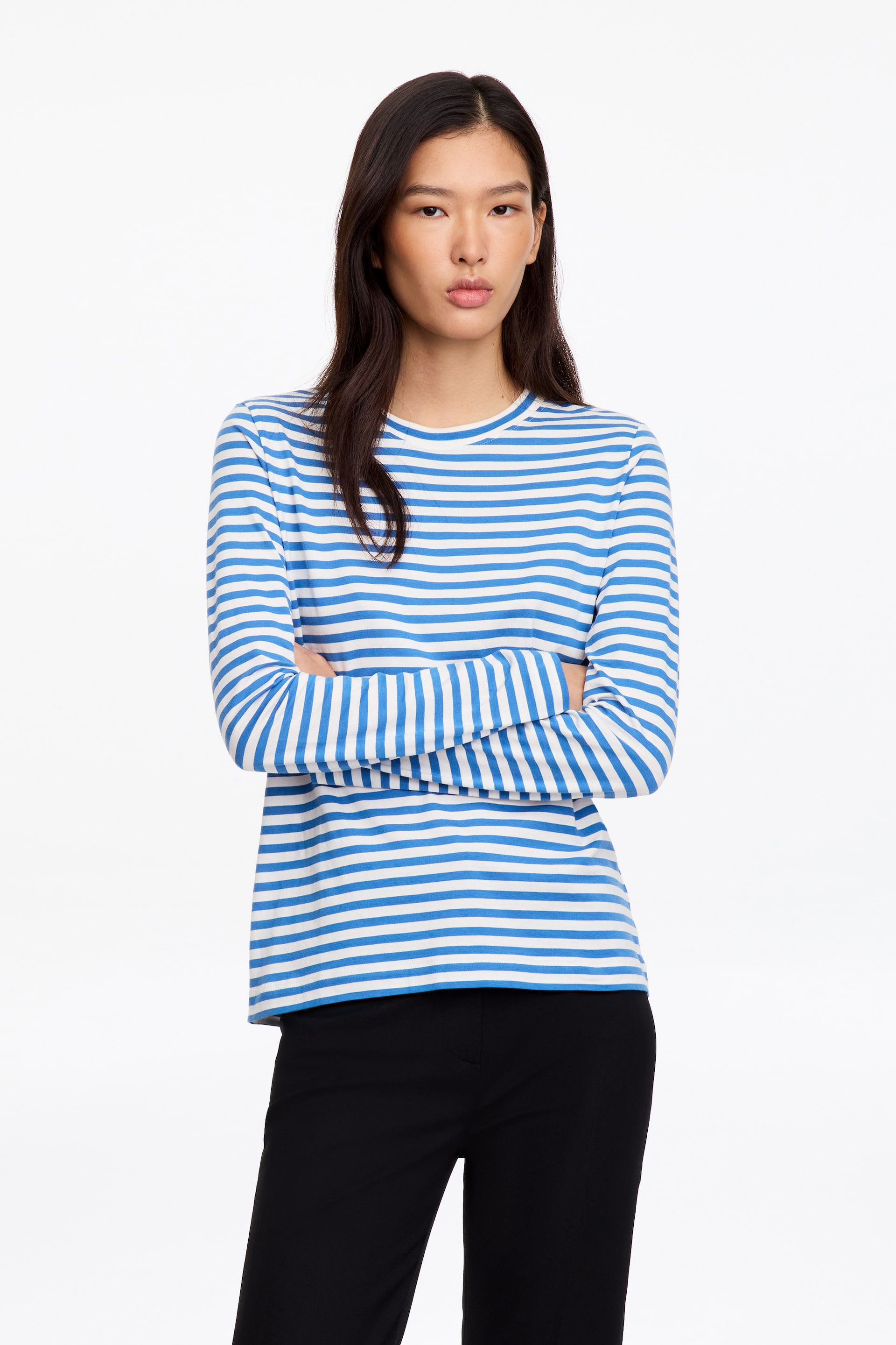 Vezi imaginea mai mare: Un tricou cu mânecă lungă și guler rotund, cu dungi orizontale alb-albastre, este purtat de model, care stă cu brațele încrucișate. Topul din bumbac, cu croială regular fit, este asortat cu pantaloni negri.
