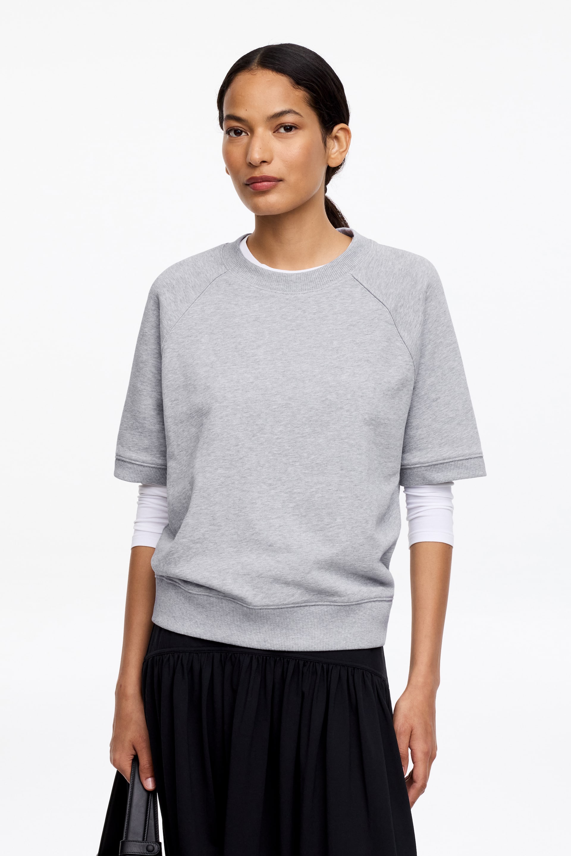 Ver imagem maior: Uma sweatshirt cinzenta de mangas curtas apresenta uma gola redonda canelada, mangas raglan, punhos canelados e uma bainha canelada. Uma camisa branca de mangas compridas está por baixo, com as suas mangas a estenderem-se além dos punhos da sweatshirt.