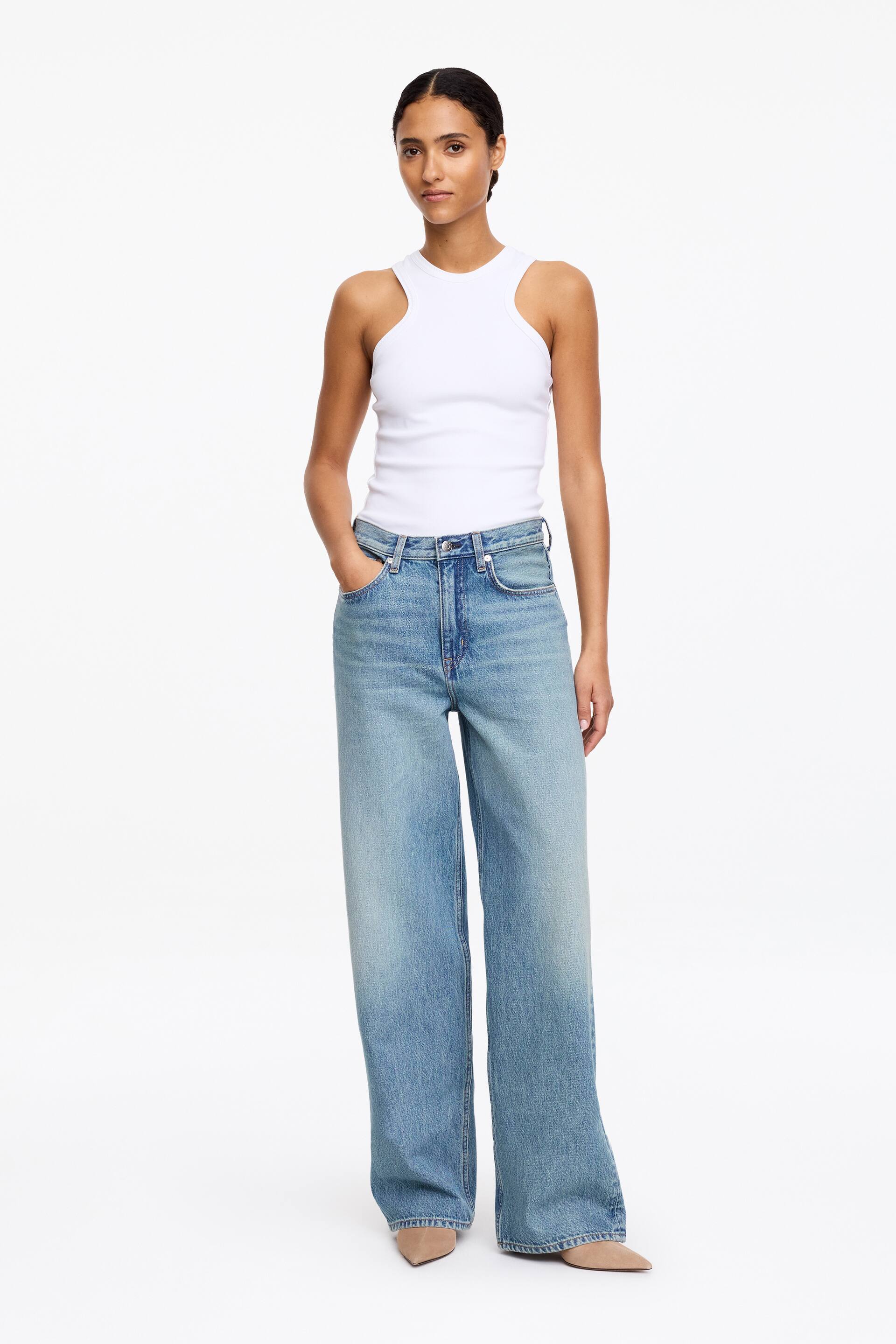 Agrandir l'image: Un débardeur blanc côtelé est porté, avec un col montant et des emmanchures découpées. Il est associé à un jean bleu clair à jambes larges, présentant un délavage dégradé et une coupe taille haute, recouvrant le haut de chaussures à bout pointu.