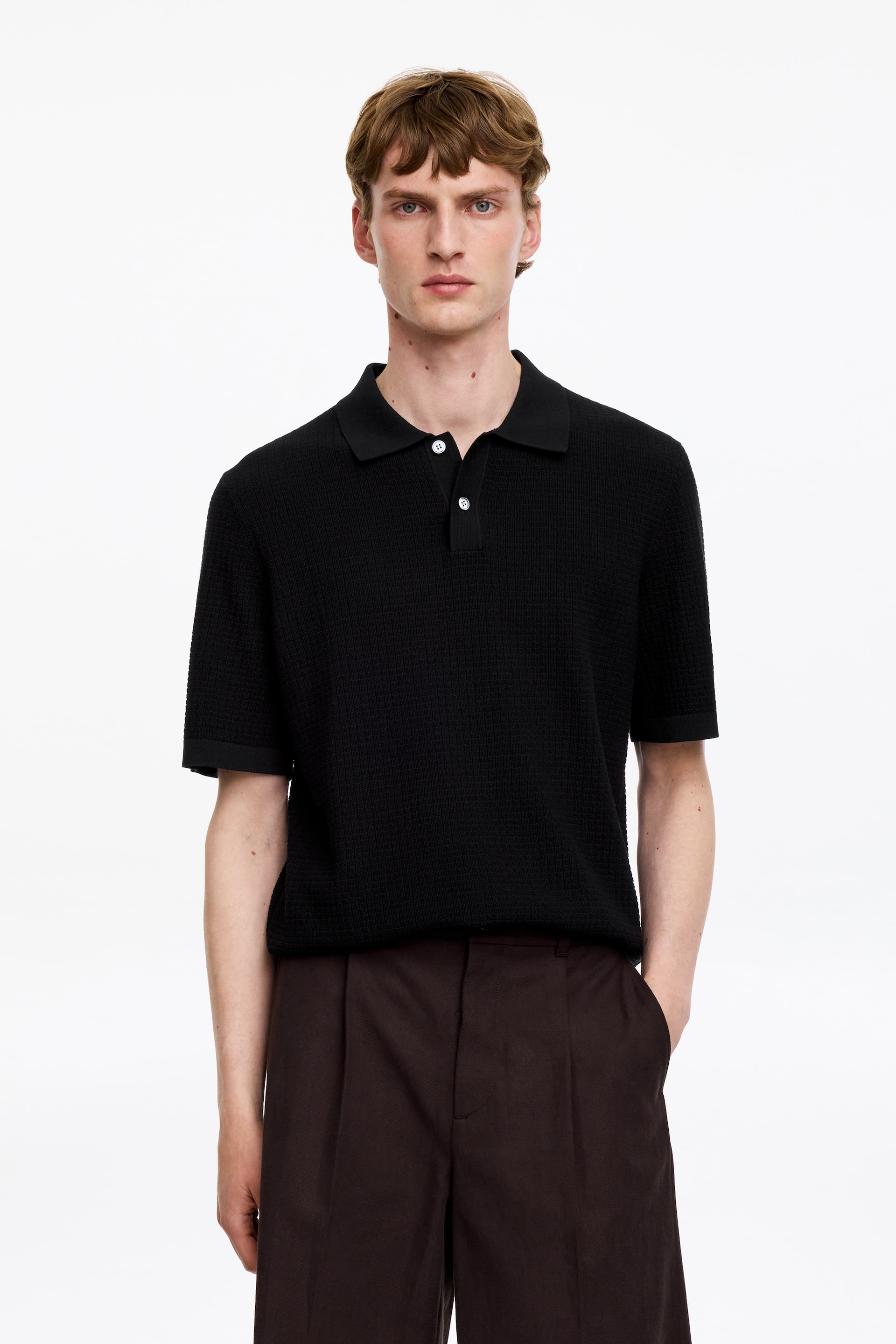 Grösseres Bild ansehen: Ein schwarzes Strick-Poloshirt mit einer strukturierten Moosstrick-Optik und kurzen Ärmeln wird vom Model getragen. Es verfügt über einen klassischen Polokragen und eine halbe Knopfleiste mit Perlmuttknöpfen.