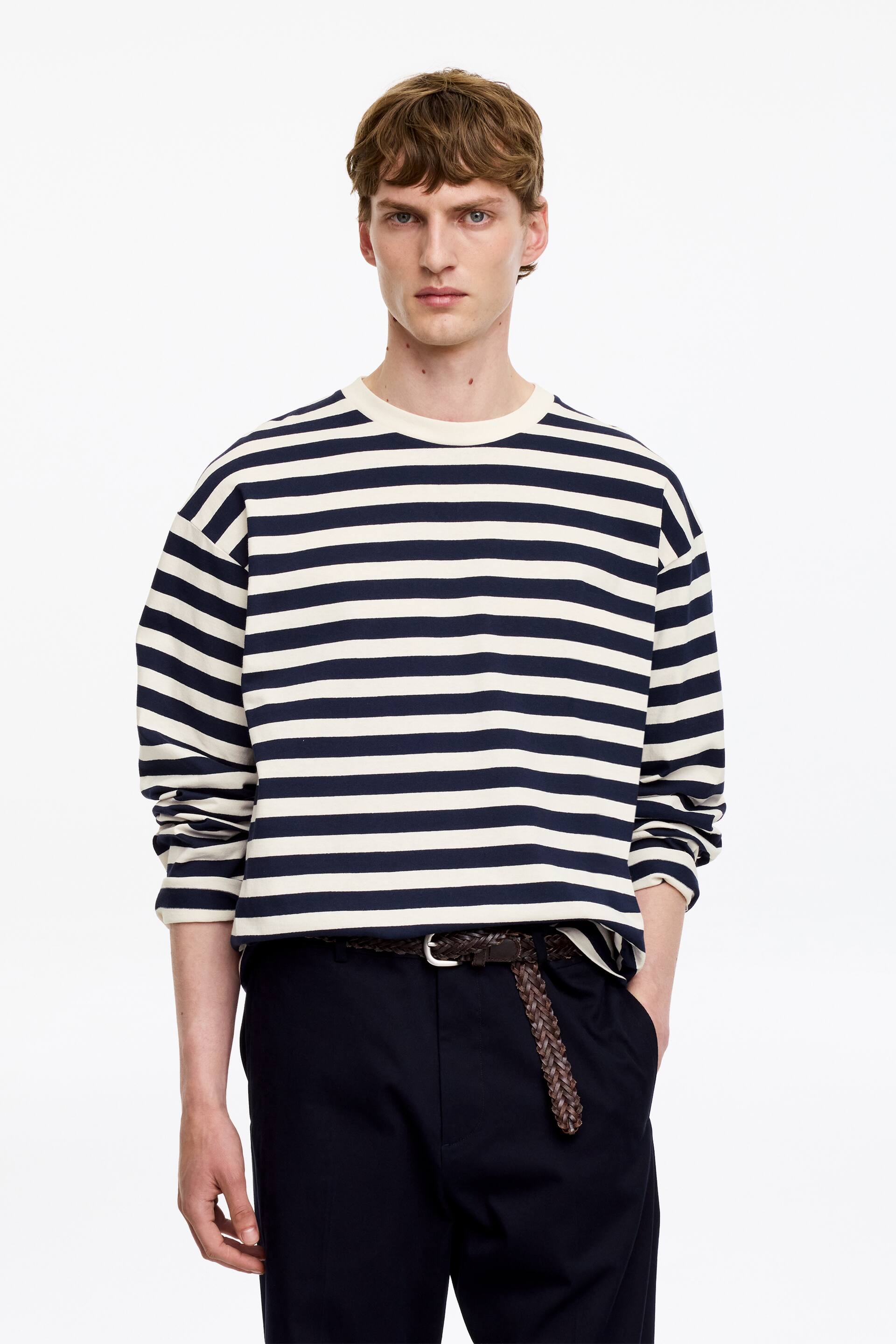 Grotere afbeelding bekijken: Een T-shirt met lange mouwen met marineblauwe en off-white strepen en een geribbelde ronde hals wordt gedragen door het model. Het T-shirt met een ontspannen pasvorm is gedeeltelijk ingestopt in een donkere broek, waarbij de hand van het model in een broekzak zit.