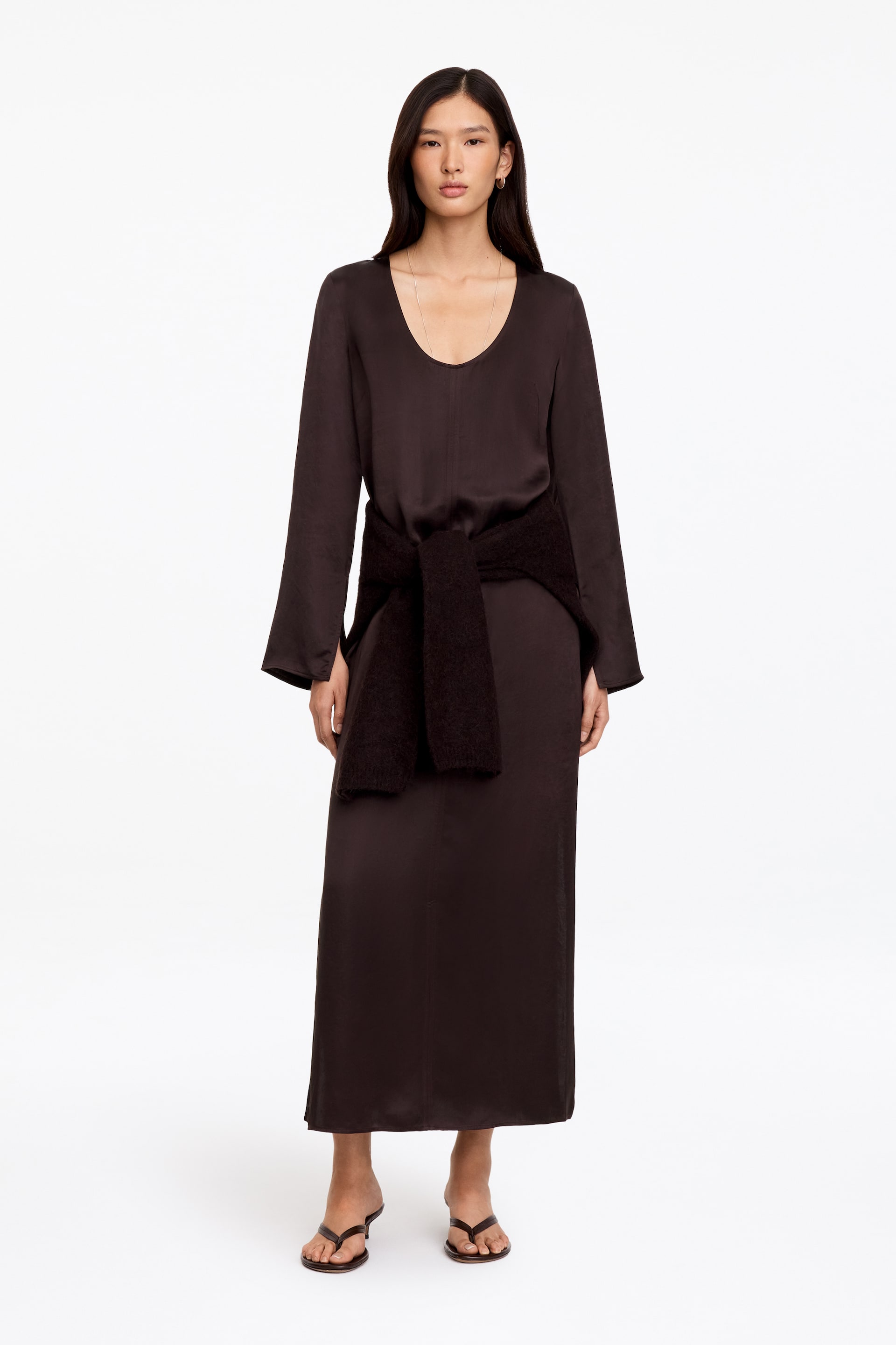 Agrandir l'image: Une robe longue en satin brun foncé, offrant un léger brillant, avec un col rond et des manches longues à poignets fendus, est portée par le mannequin. Un pull en maille foncé est stylé en étant noué autour de la taille de la robe.