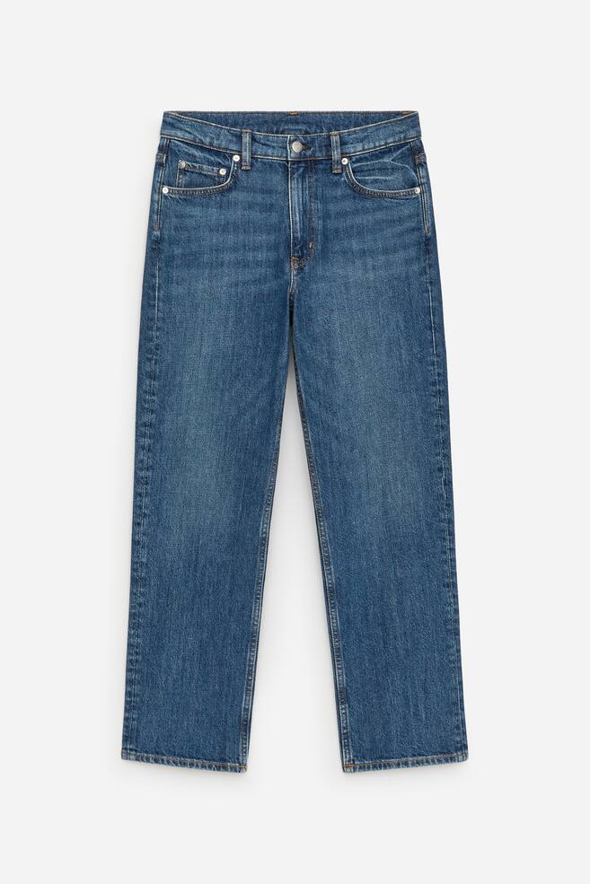 O pereche de blugi din denim albastru își prezintă partea FRONTALĂ, având o talie cu găici pentru curea, o închidere cu nasture și fermoar, și buzunare frontale cu un buzunar pentru monede. Blugii prezintă un croi conic, cu picior scurtat. - 1