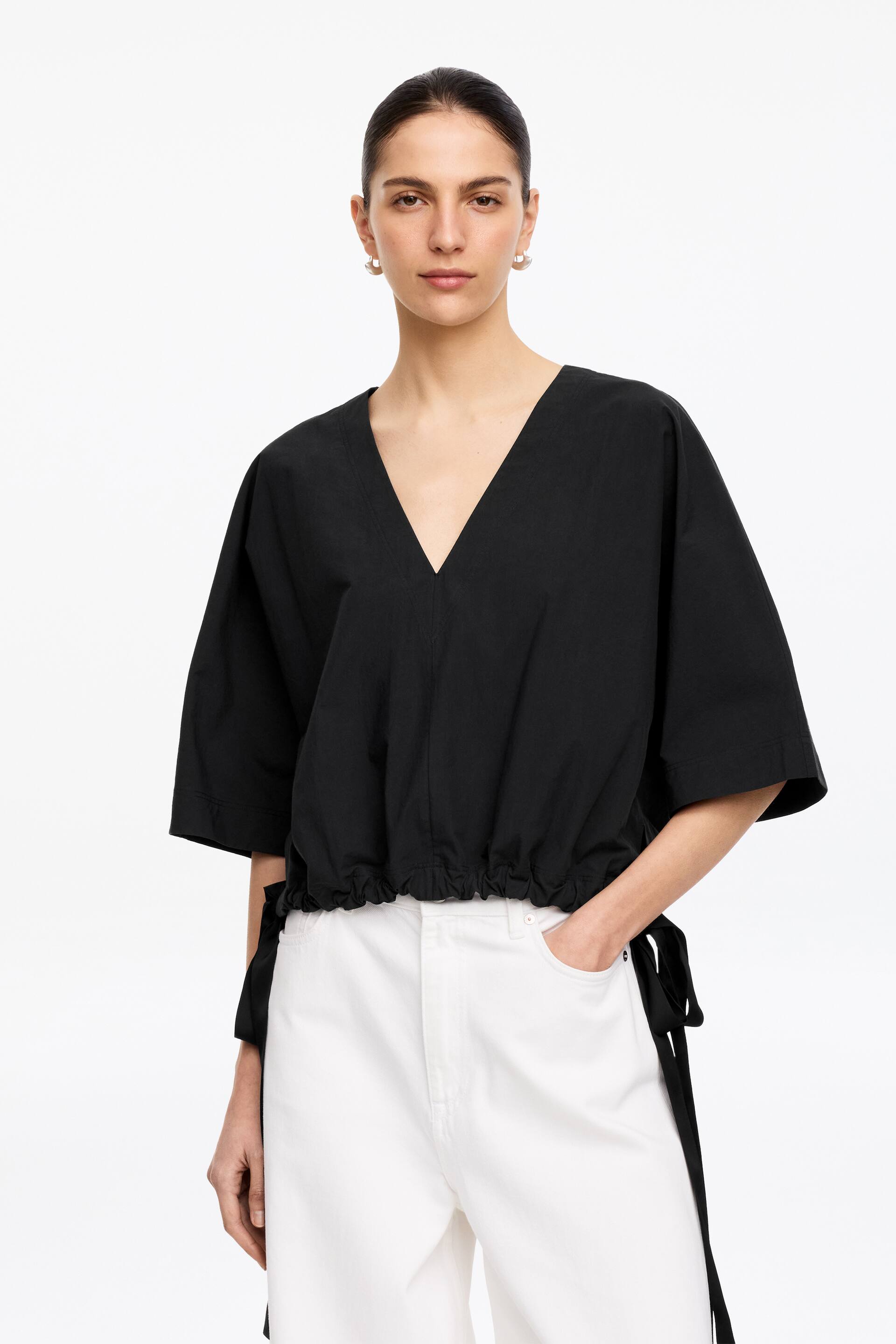View larger image: A black V-neck top featuring wide, elbow-length sleeves. It showcases a cinched waist with visible ties hanging down the sides, creating a gathered effect.