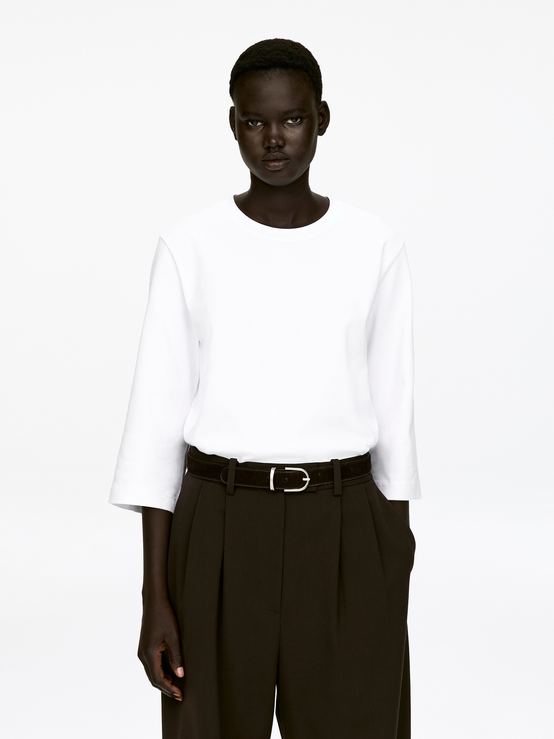 Ver imagen más grande: Una camiseta blanca de cuello redondo con mangas de 3/4, que presenta un aspecto suave y estructurado, está siendo llevada por el modelo. La camiseta está metida dentro de unos pantalones oscuros de pinzas, que están asegurados con un cinturón oscuro.