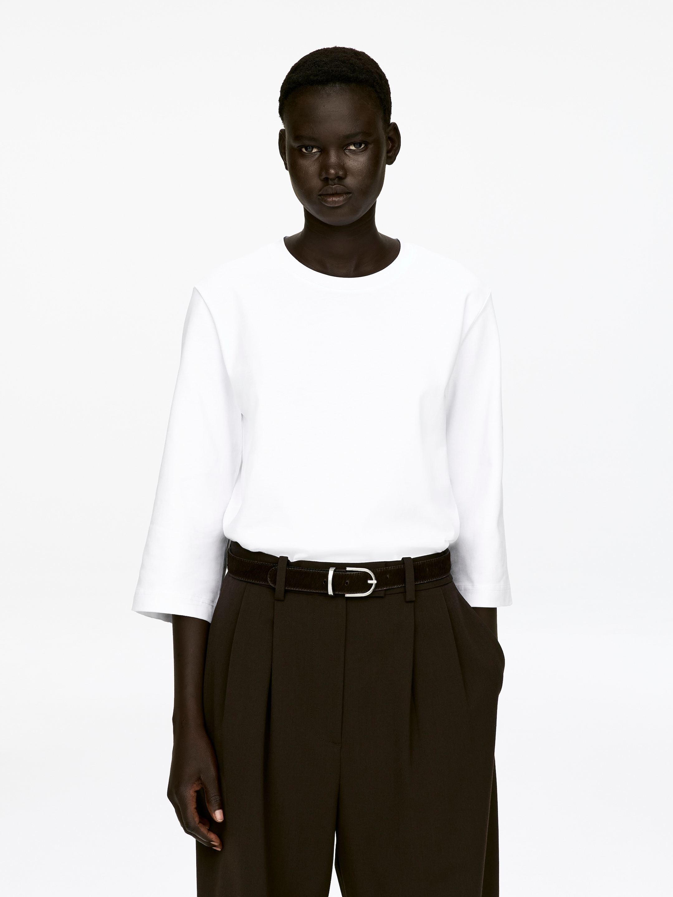Agrandir l'image: Un T-shirt blanc à col rond avec des manches 3/4, présentant une apparence lisse et structurée, est porté par le mannequin. Le T-shirt est rentré dans un pantalon plissé foncé, qui est maintenu en place par une ceinture foncée.