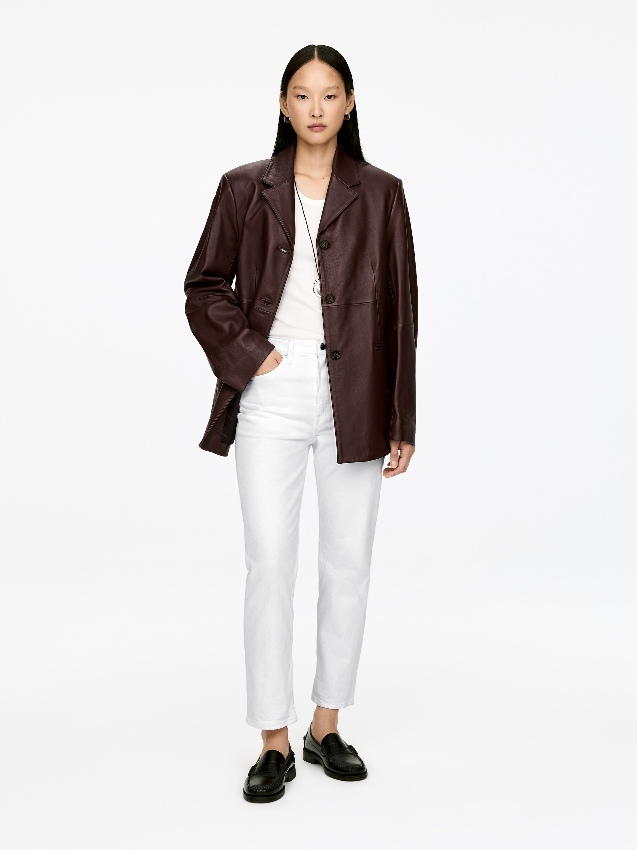 Agrandir l'image: Une veste en cuir brun foncé fait face à l'avant, avec un col à revers et trois boutons foncés sur le devant. Elle présente une coupe surdimensionnée, portée ouverte sur un t-shirt blanc et un jean blanc.