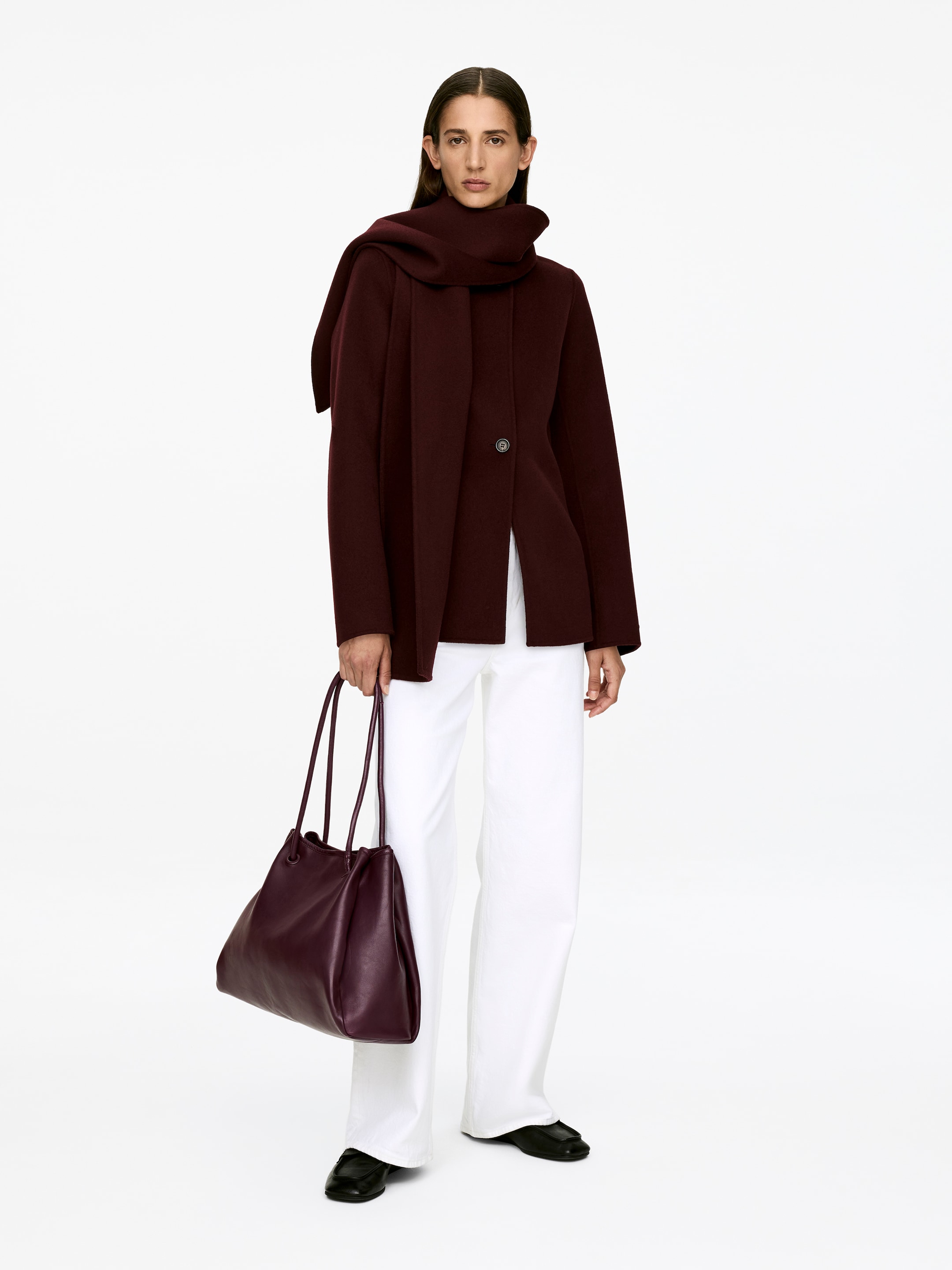 Ver imagem maior: Mala de Couro tipo Tote. Uma mala tipo tote de couro em tom de ameixa, com um topo franzido e duas alças finas, está a ser carregada pela modelo. A mala macia e flexível está posicionada ao lado dela.