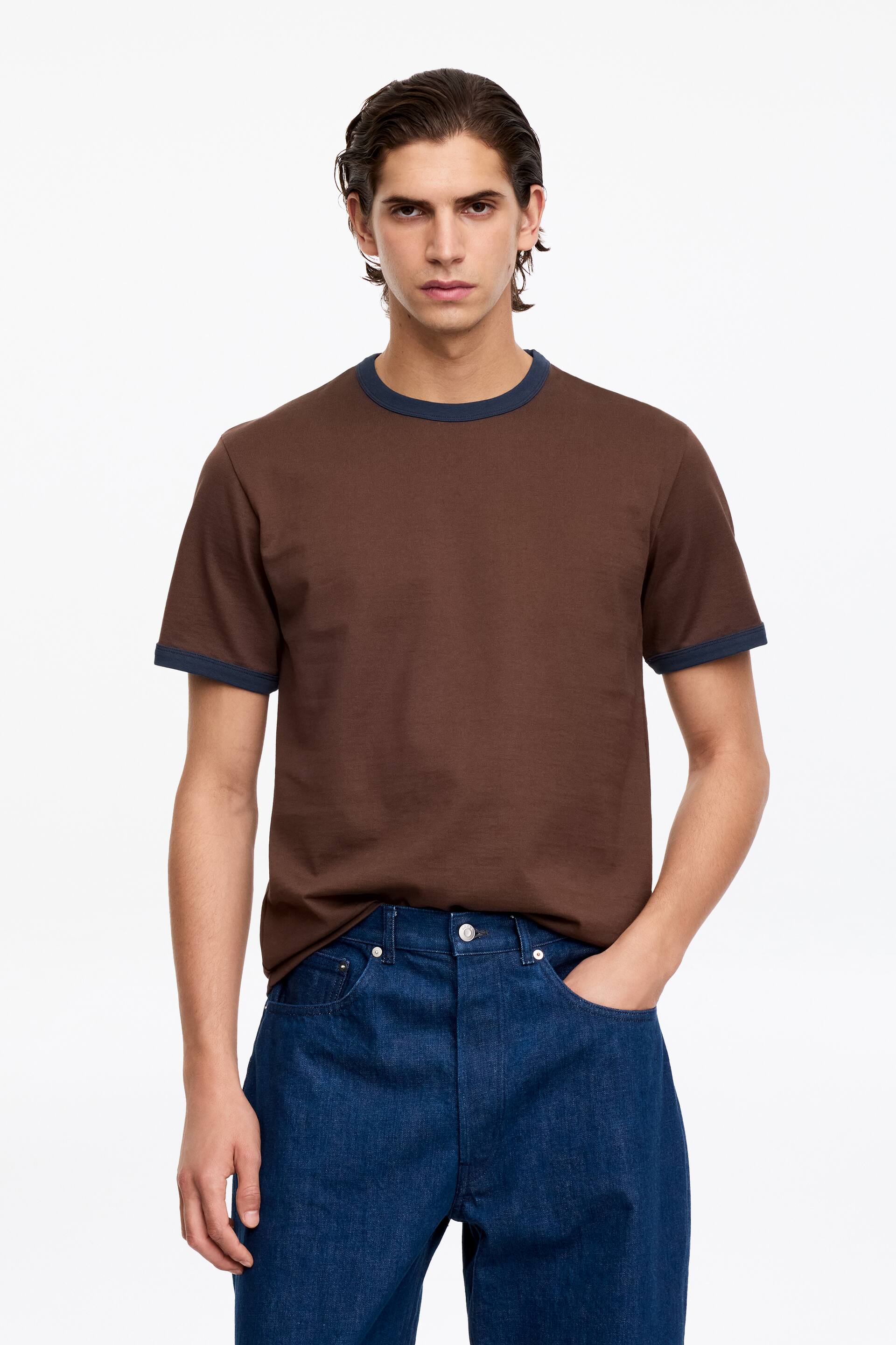 Größeres Bild anzeigen: Ein braunes Ringer-T-Shirt wird getragen, mit einem marineblauen Rundhalsausschnitt und passenden marineblauen Abschlüssen an den kurzen Ärmeln. Die rechte untere Seite des T-Shirts ist eingesteckt, während die linke Seite locker getragen wird.