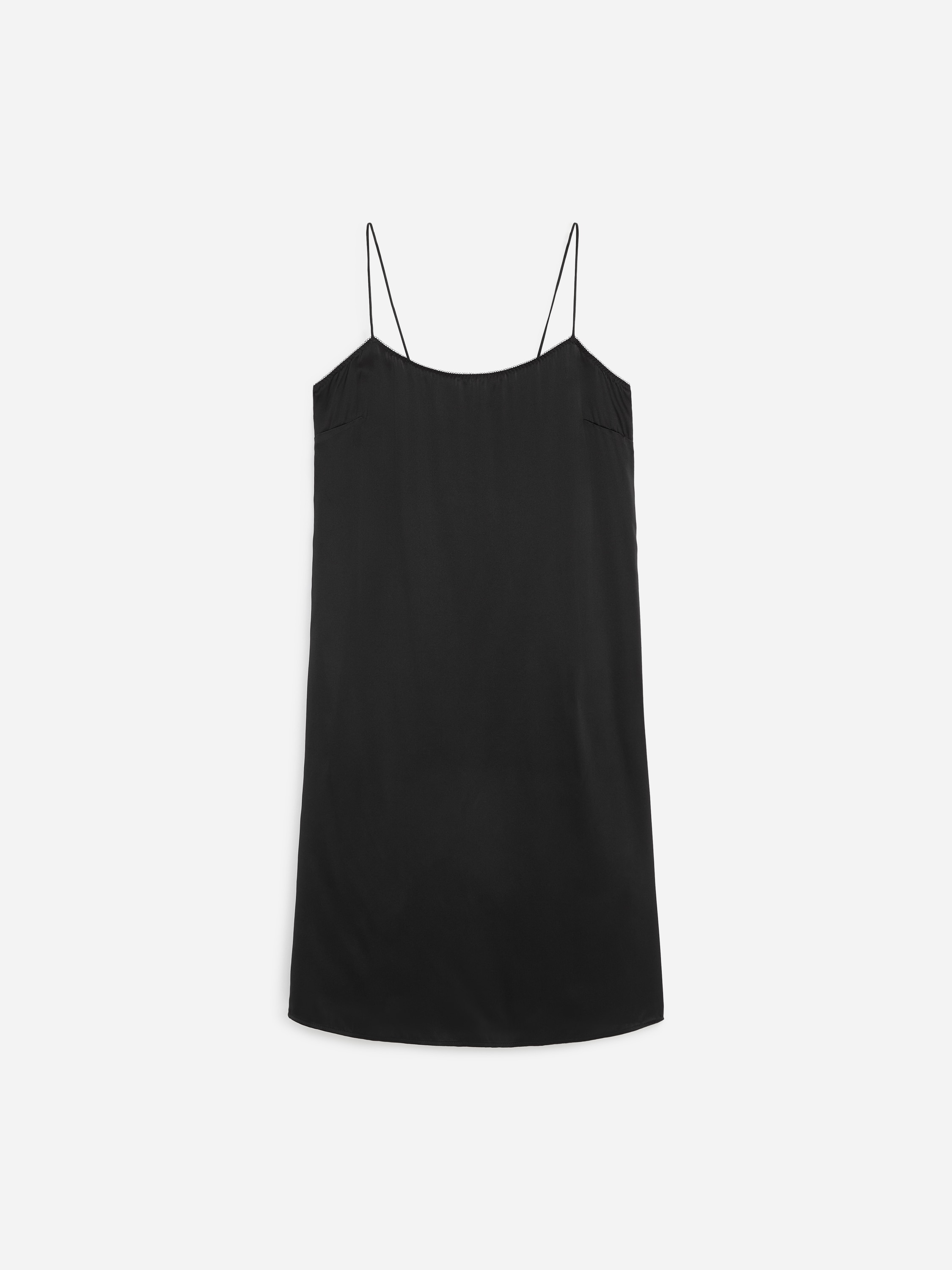 A black midi slip dress with a straight neckline, delicate shoulder straps and a relaxed silhouette is being worn by the model. The dress falls below the knee and is styled with black flat thong sandals.