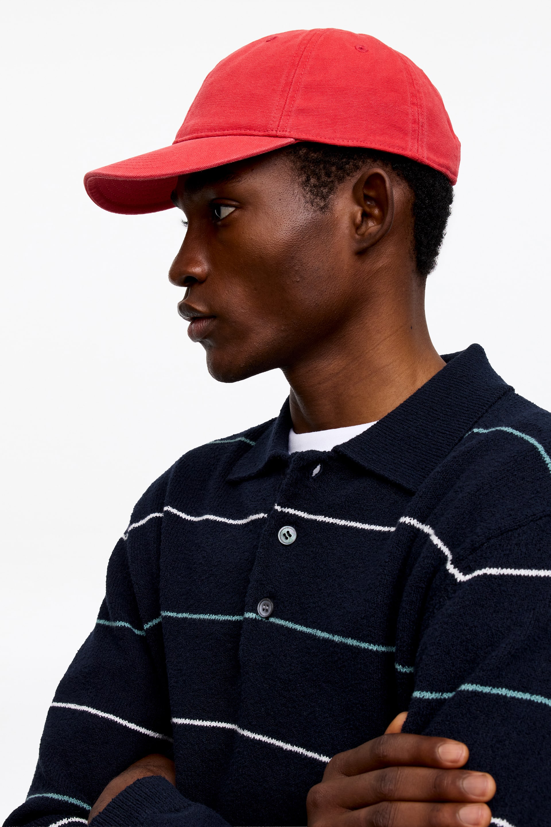Visualizza immagine più grande: Un berretto da baseball rosso è indossato, rivolto a sinistra, con una visiera curva e un tessuto strutturato. Una polo in maglia blu navy è indossata, con strisce orizzontali bianche e azzurro chiaro, un colletto aperto e un'allacciatura a due bottoni con bottoni scuri, sovrapposta a una t-shirt bianca.