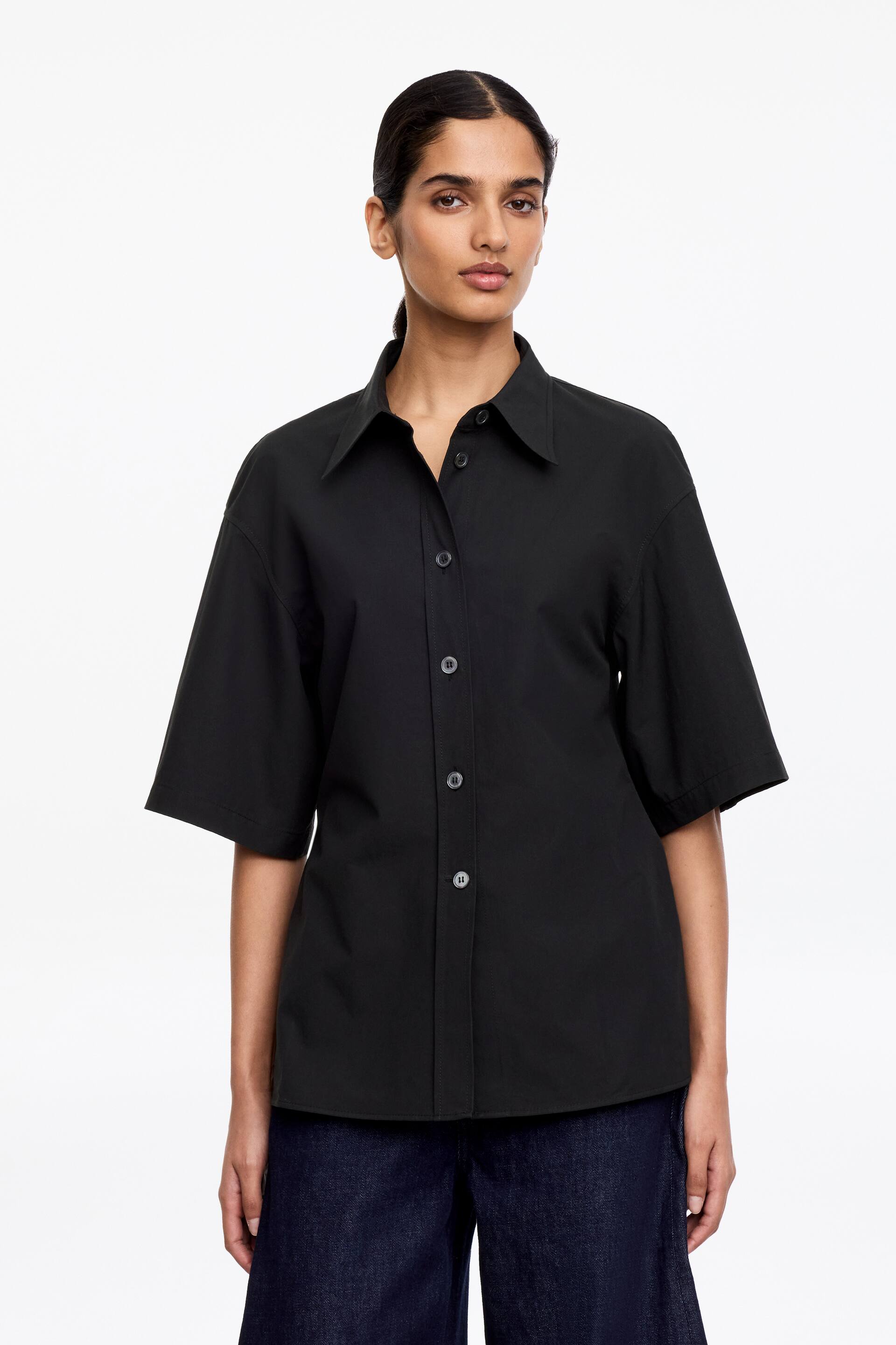 Grotere afbeelding bekijken: Een zwarte korte mouwen overhemd wordt gepresenteerd, met een klassieke puntkraag, een volledige knoopsluiting aan de voorkant met donkere knopen, en brede, ellebooglange mouwen met een ontspannen pasvorm.