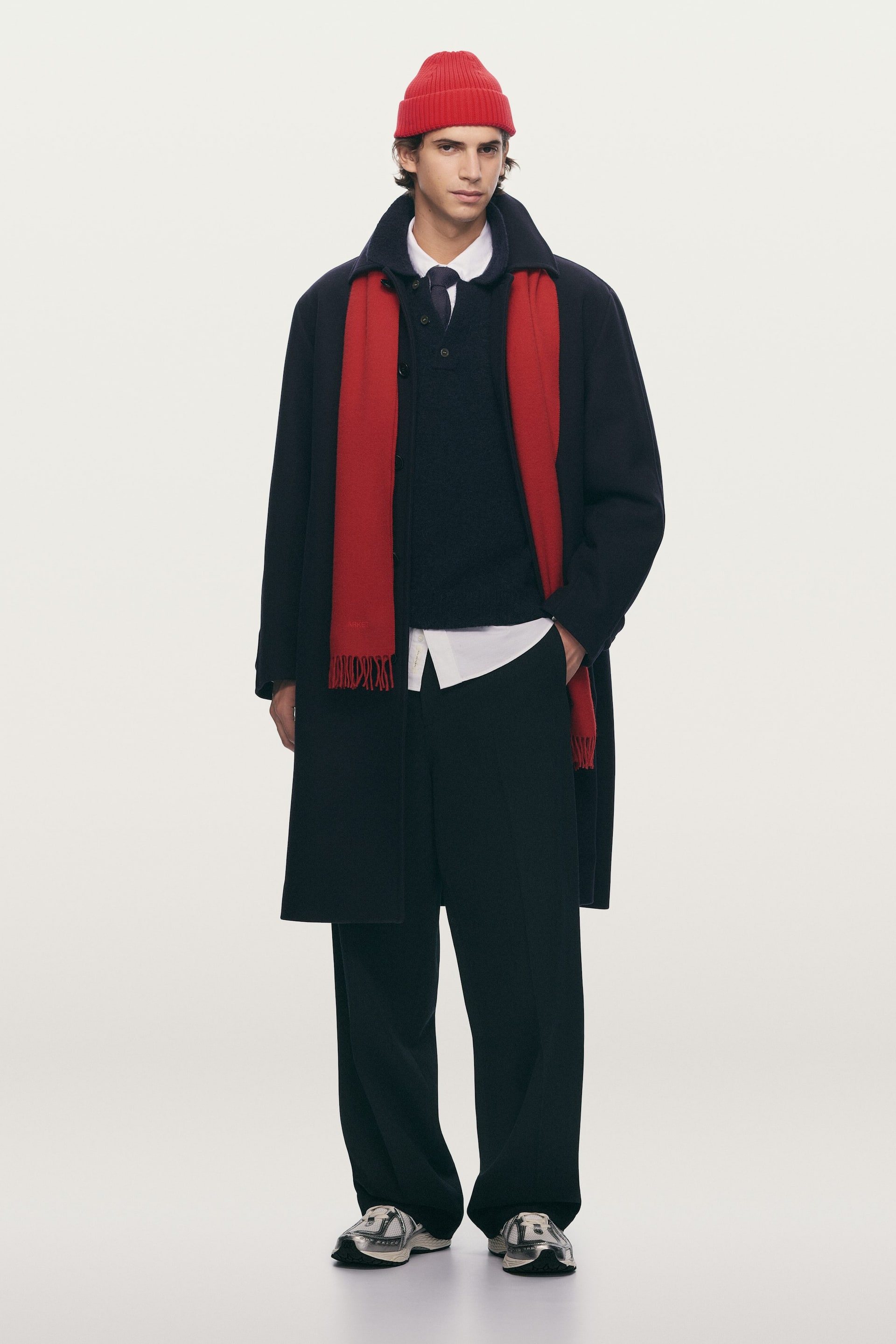 Se større billede: Et fuldt outfit præsenterer en marineblå lang frakke, båret åben og fremadvendt, lagdelt over et rødt tørklæde draperet rundt om halsen. Underneden bæres en marineblå henley-stil sweater over en hvid skjorte med krave og marineblåt slips, kombineret med mørke, løstsiddende bukser, flerfarvede sneakers og en rød ribstrikket hue.