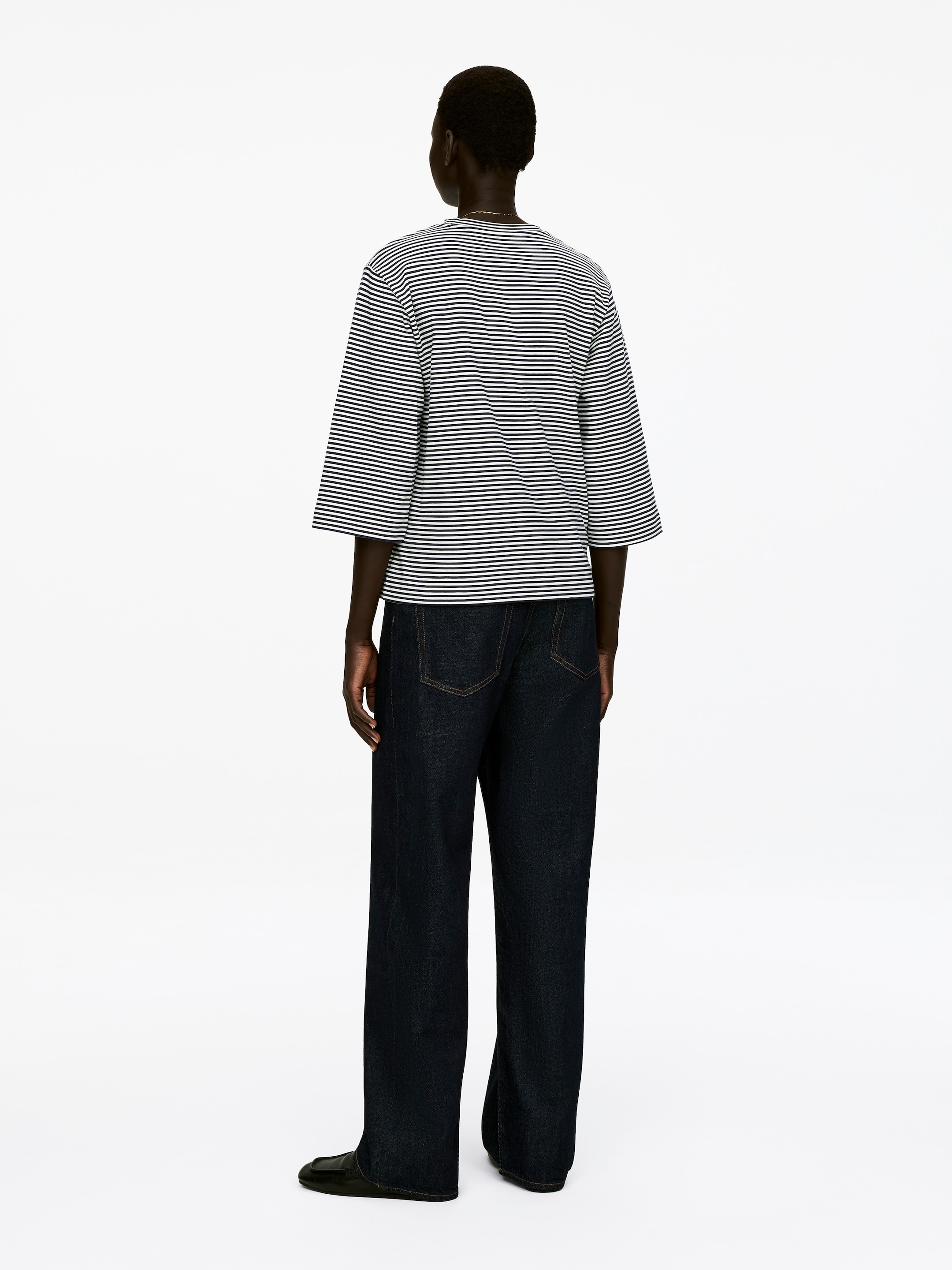 Agrandir l'image: Un t-shirt à col rond, réalisé en jersey de coton épais et frais avec des rayures horizontales noires et blanches et des manches 3/4, est porté par le mannequin. Il est assorti d'un pantalon à larges jambes de lavage foncé et de chaussures plates sombres.