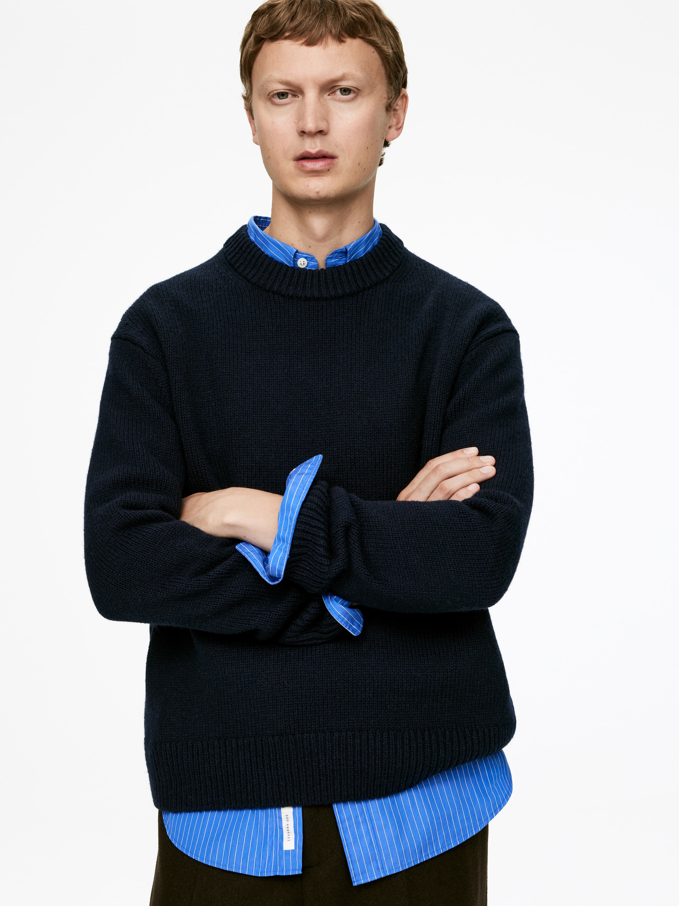 View larger image: A navy blue crew neck sweater is being presented front-facing, featuring a relaxed fit and a visible ribbed knit texture. It is layered over a blue and white striped collared shirt, with the sweater's ribbed cuffs and hem being clearly visible.