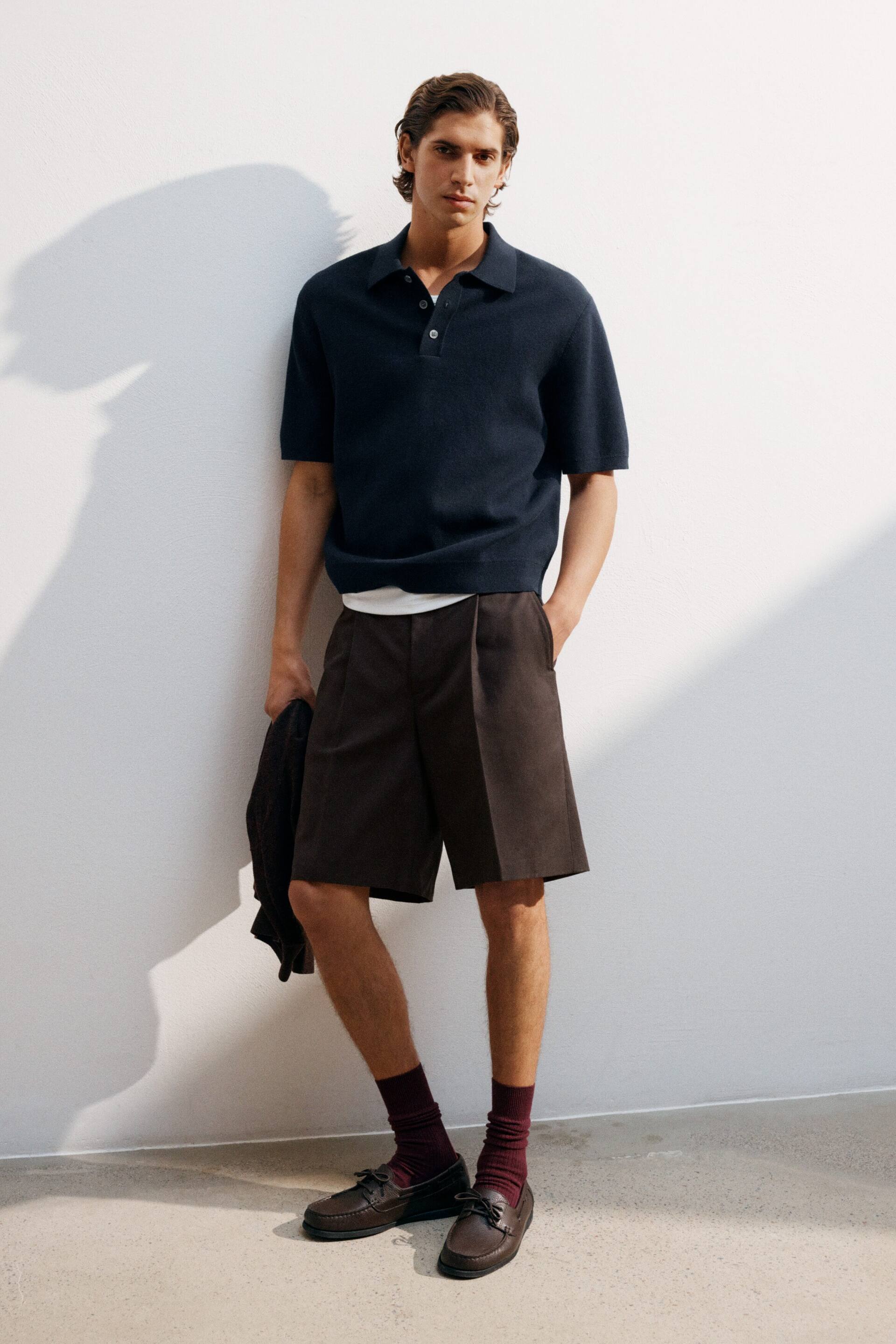 Grösseres Bild ansehen: Ein dunkelblaues Strick-Poloshirt wird getragen, das mit einem spitzen Kragen, einer Drei-Knopf-Leiste mit weißen Knöpfen und kurzen Ärmeln ausgestattet ist. Es zeigt eine lockere Passform und einen gerippten Saum, gestylt über einem weißen T-Shirt und in dunkelbraune Shorts gesteckt.