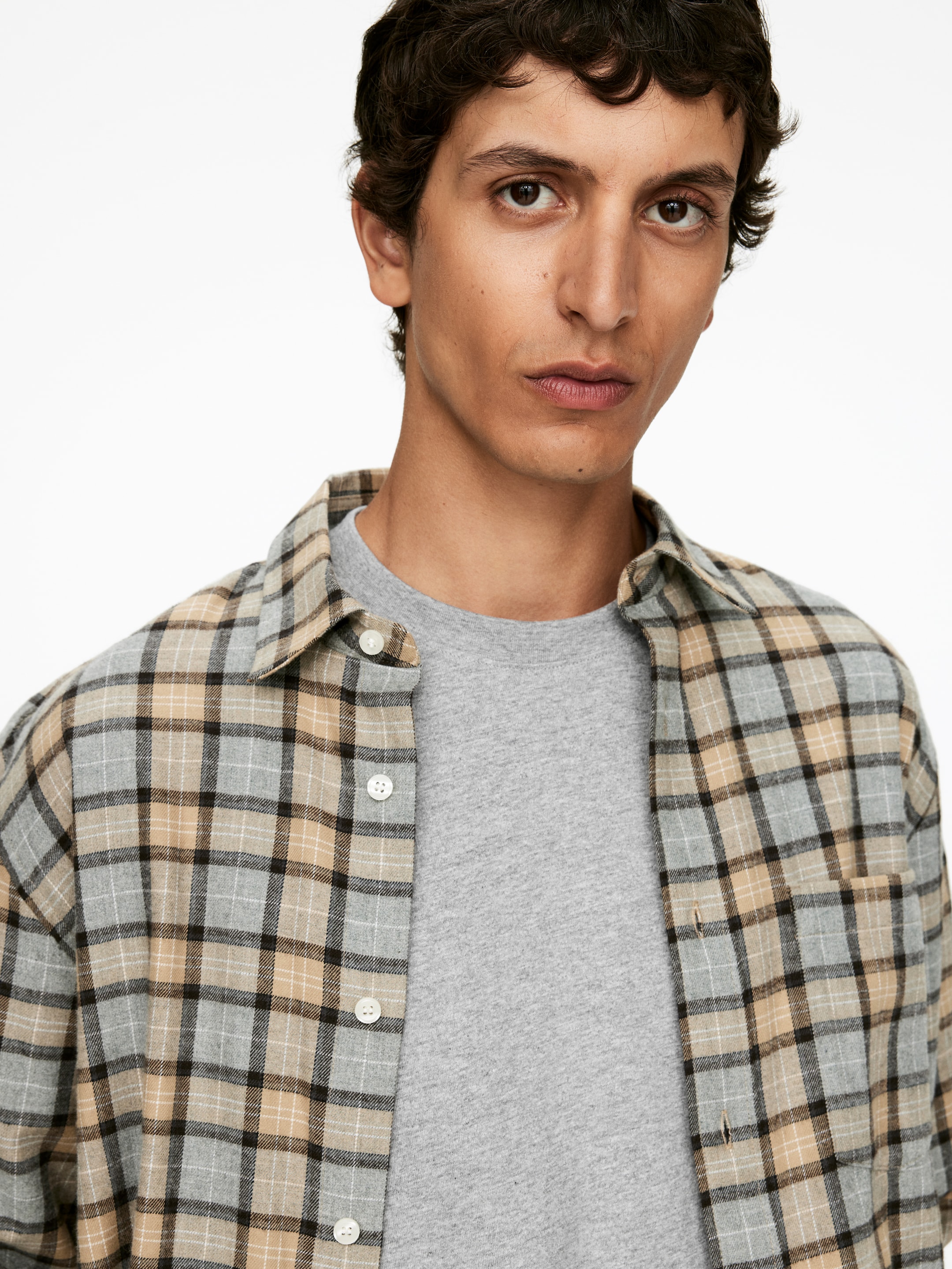 Visualizza immagine più grande: Una camicia a quadri grigia chiara, beige e nera è indossata sbottonata sopra una t-shirt grigia chiara con scollo a girocollo. La camicia presenta bottoni bianchi e una singola tasca sul petto, ed è rivolta in avanti.