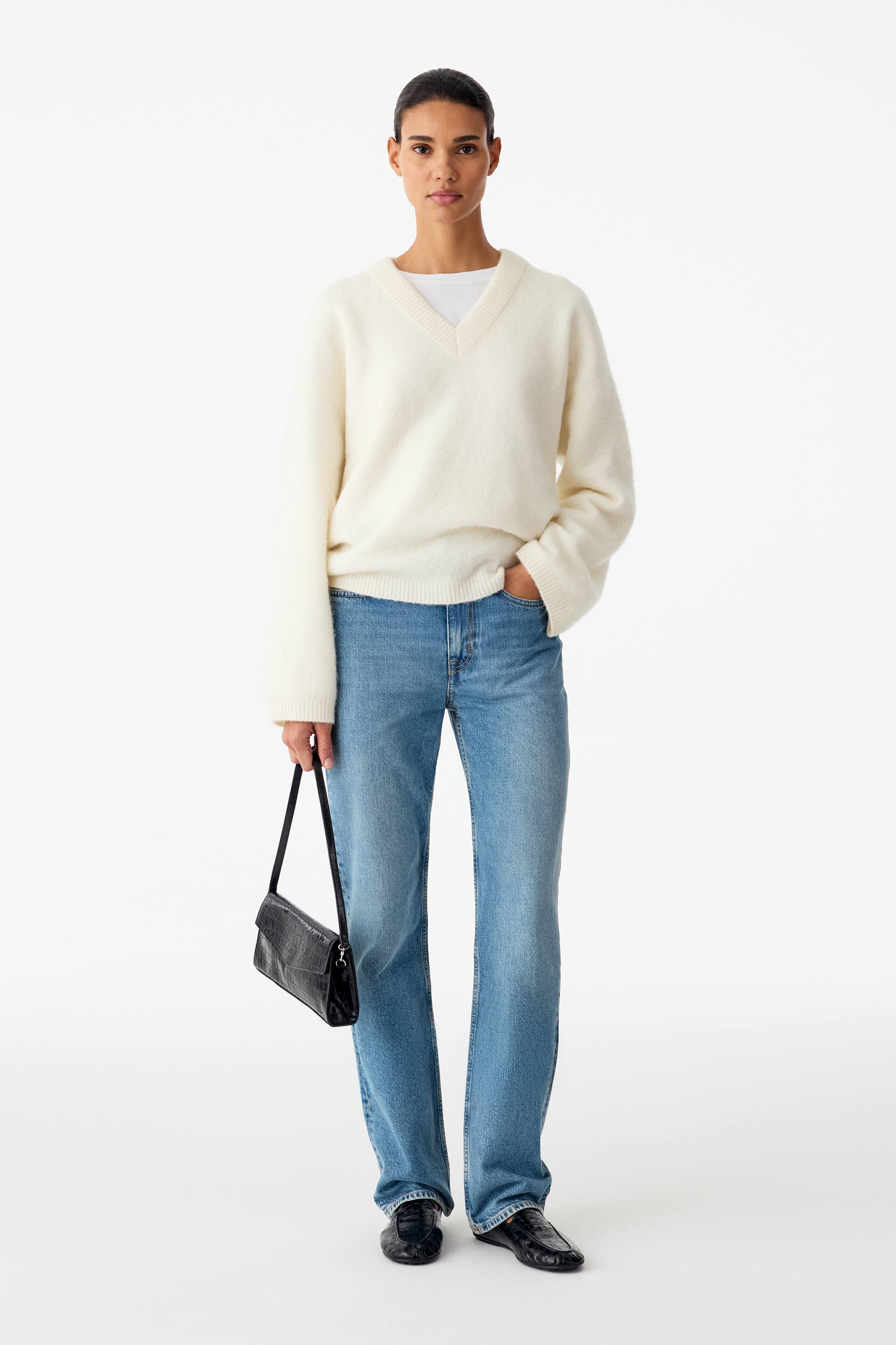 Visa större bild: Ljus tvätt, raka denimjeans med medelhög midja och smal passform bärs av modellen, som vilar ena handen i en ficka. Jeansen kombineras med en krämfärgad v-ringad tröja, en vit t-shirt, svarta ballerinaskor och en svart axelväska.