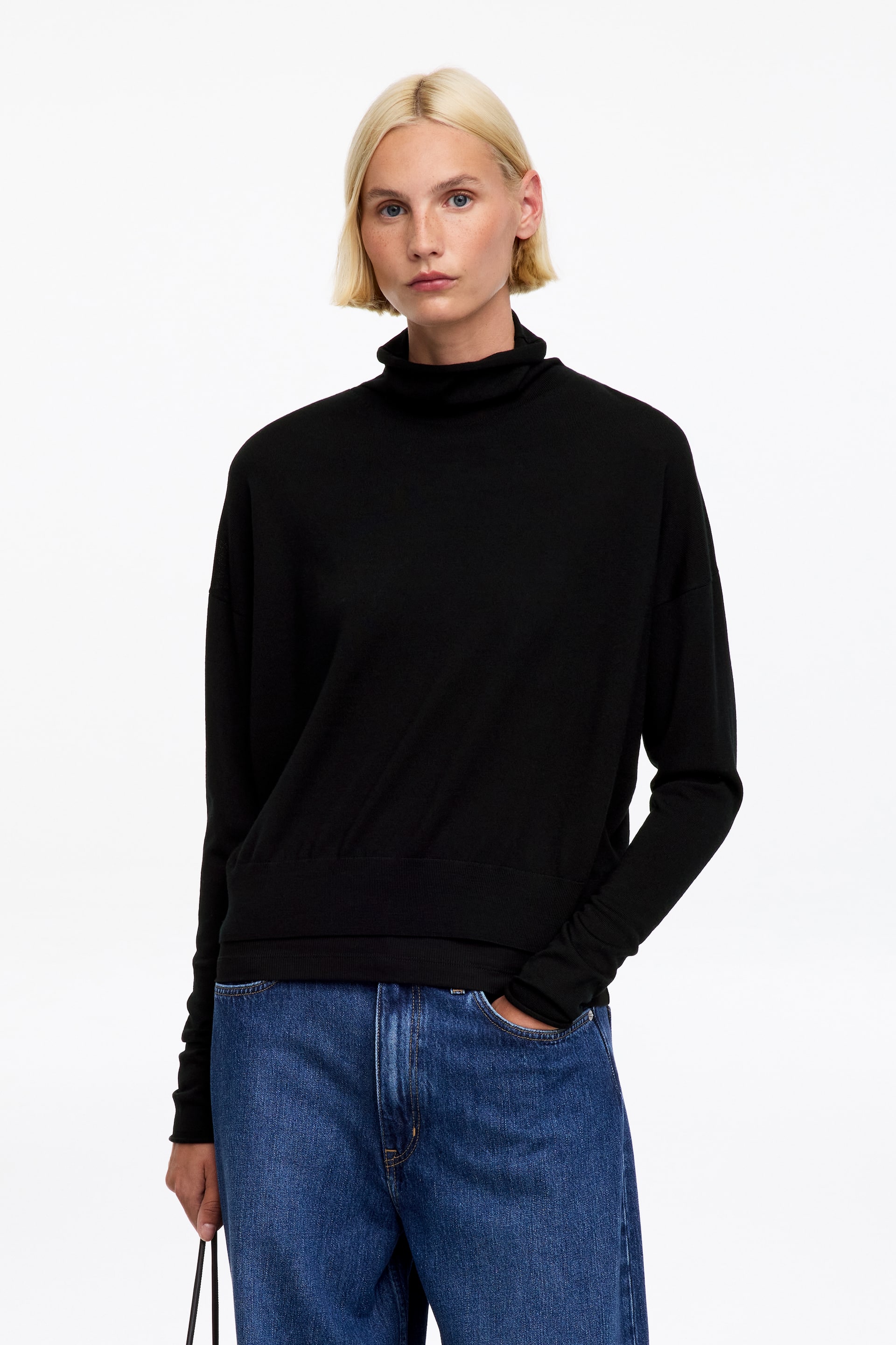 Visualizza immagine più grande: Il modello indossa un maglione nero a collo alto. Questo maglione dalla vestibilità rilassata presenta un collo alto, maniche lunghe con polsini a coste e fondo a coste. Il modello sta infilando una mano in una tasca.