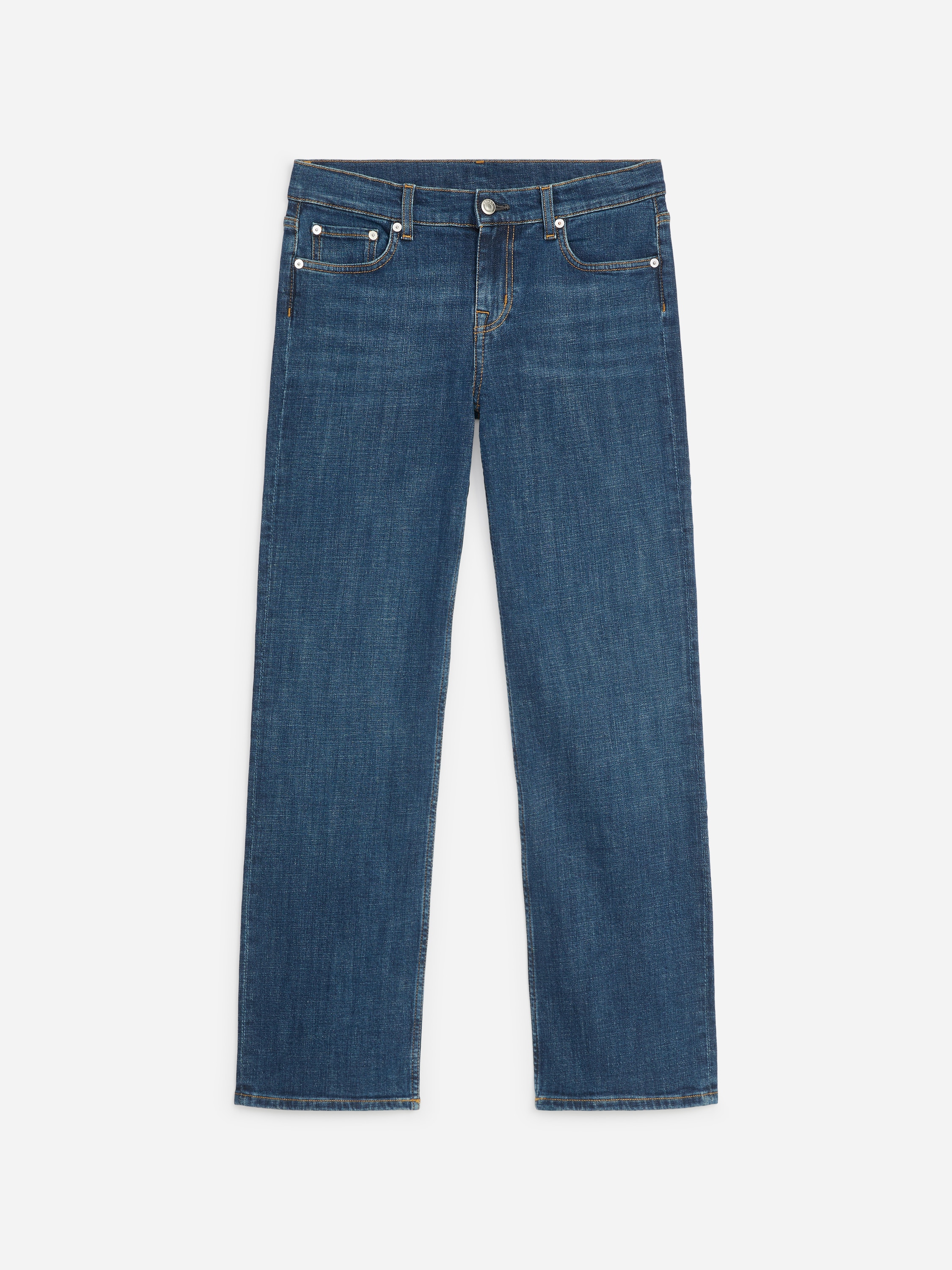 Unos vaqueros de denim con lavado azul oscuro, de tiro bajo, corte slim y pernera recta, lucidos por la modelo. Los vaqueros se combinan con una camisa blanca de manga larga metida por dentro y zapatos negros.
