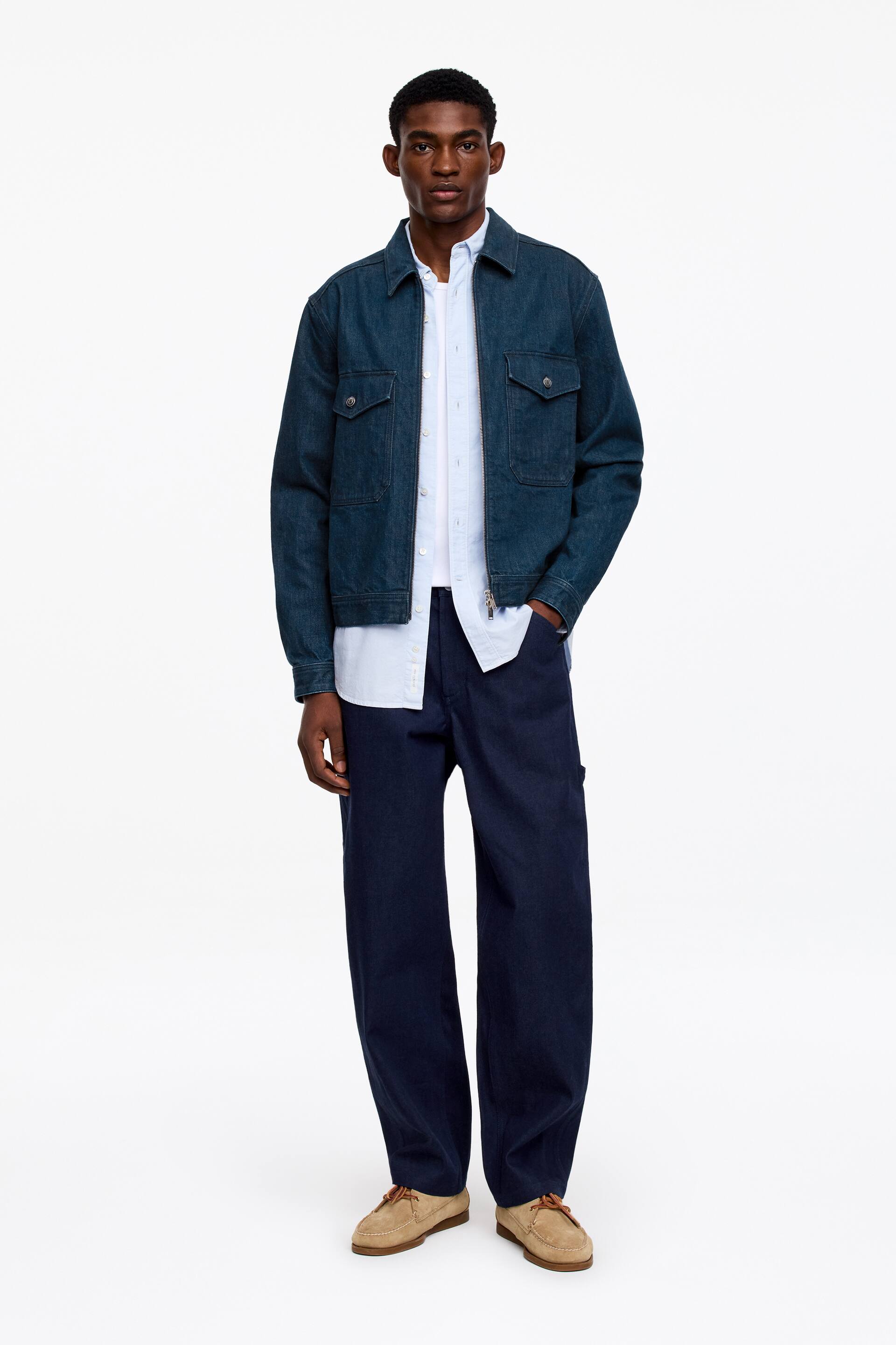Visa större bild: En mörkblå denimjacka presenteras från en framåtvänd vinkel, med en klassisk krage, en hel dragkedjestängning, två bröstfickor med knäppflikar och två nedre påsydda fickor. Den bärs uppknäppt över en ljusblå mönstrad skjorta med knappar, tillsammans med mörkblå vida arbetsbyxor och bruna mockaloafers som kompletterar helheten.