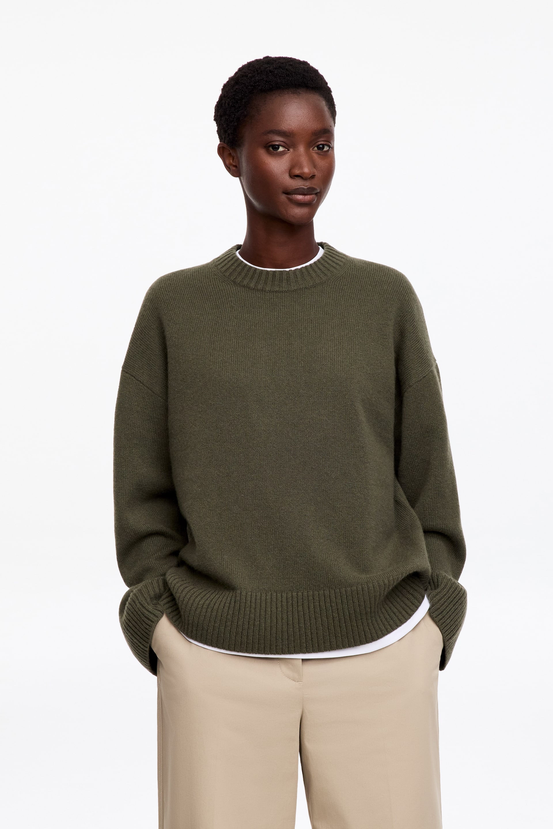 Se større billede: En grøn striktrøje med rund hals, sænkede skuldre og ribkanter bæres af modellen over en lagdelt hvid T-shirt. Den løstsiddende sweater styles med beige bukser, mens modellen hviler hænderne i buksernes lommer.