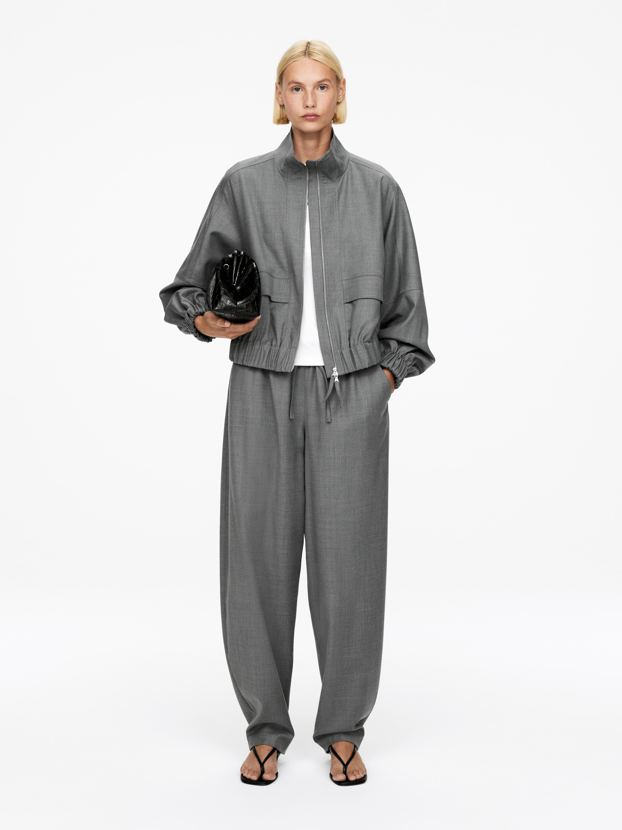 Grotere afbeelding bekijken: Een grijs gecoördineerde set wordt gepresenteerd, met een relaxed-fit jas en wijde broek. De jas is ontworpen met een opstaande kraag, een ritssluiting over de volledige lengte, twee voorflapzakken en een elastische zoom en manchetten, terwijl hij over een wit t-shirt wordt gedragen; de bijpassende wijde broek heeft een losse pasvorm en een zichtbare trekkoord taille.
