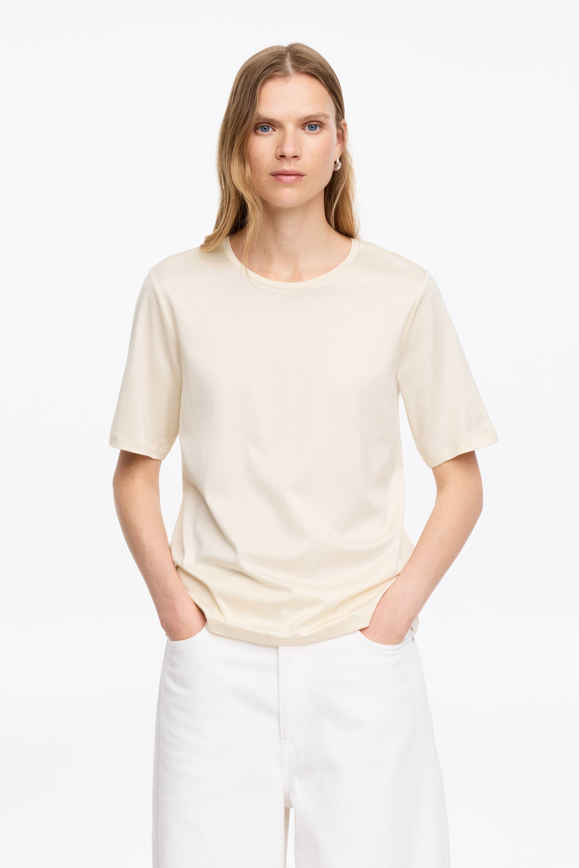 Visualizza l'immagine ingrandita: Una t-shirt color crema è indossata, con un girocollo e maniche corte. Presenta una vestibilità rilassata, cadendo dolcemente sul busto.