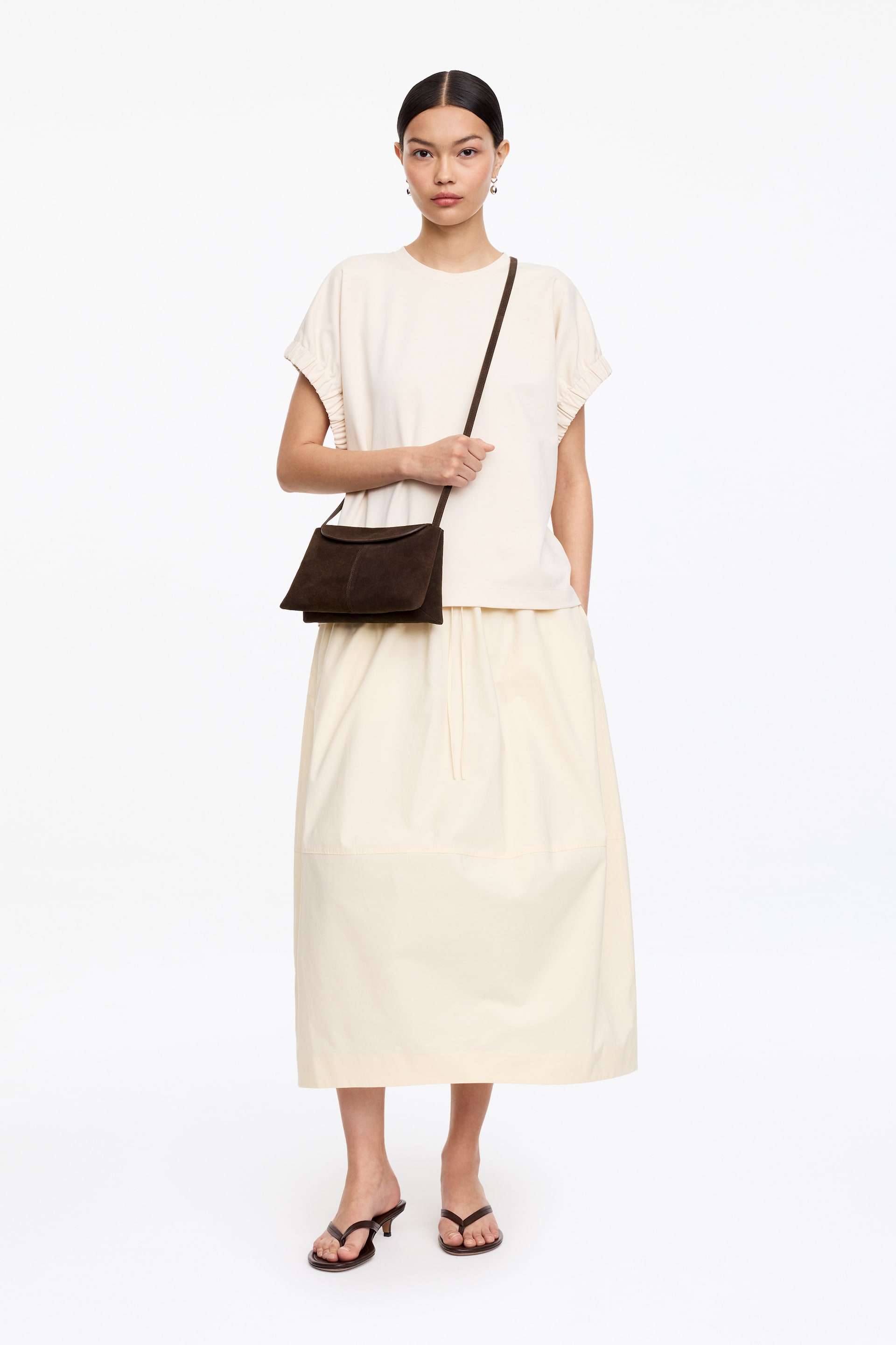 Visualizza immagine più grande: Una borsa a tracolla in pelle scamosciata marrone scuro presenta una forma rettangolare con una chiusura a patta morbida. Il suo sottile cinturino attraversa il corpo e si appoggia all'anca di chi la indossa, mentre la borsa è leggermente inclinata in avanti.