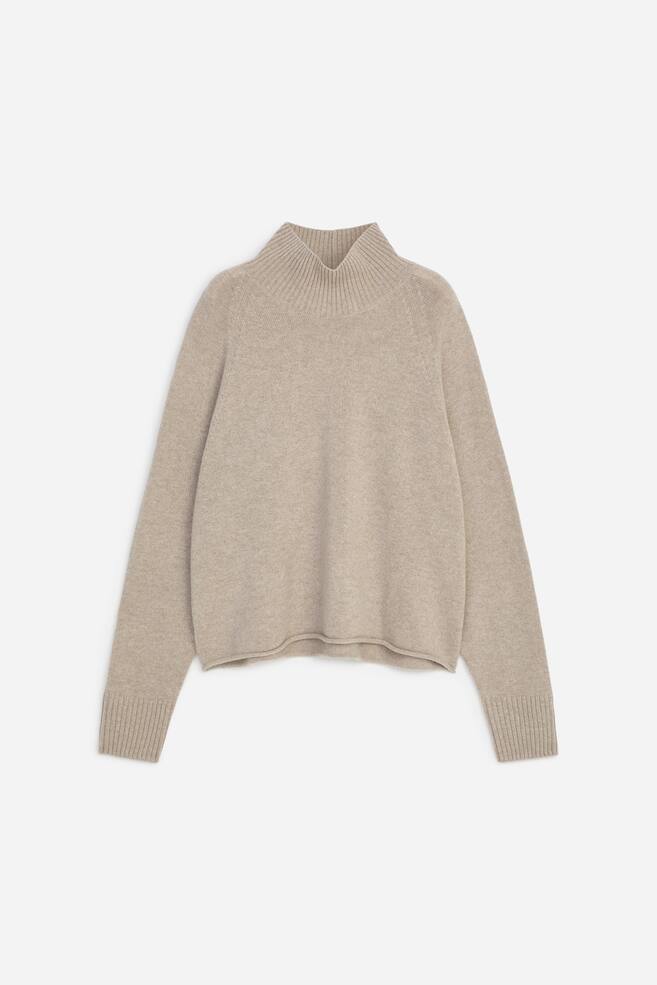 A beige knitted jumper is shown from the front, featuring a high ribbed mock neck, long raglan sleeves with wide ribbed cuffs, and a relaxed body with a subtly curved, clean hem. - 1
