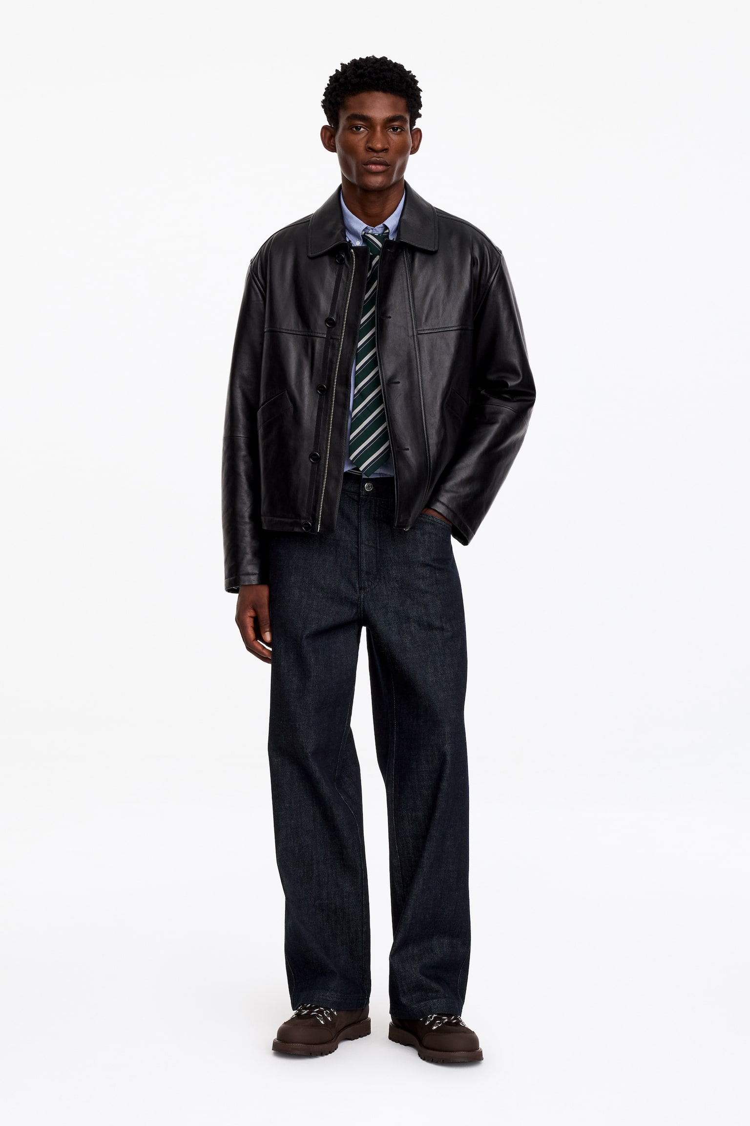 Donker gewassen denimjeans met wide-leg silhouet worden gedragen door het model, dat poseert met Ã©Ã©n hand in een zak. De jeans zijn gestyled met een zwarte leren jas, een lichtblauw overhemd met knoopsluiting, een gestreepte stropdas en bruine laarzen. - 1