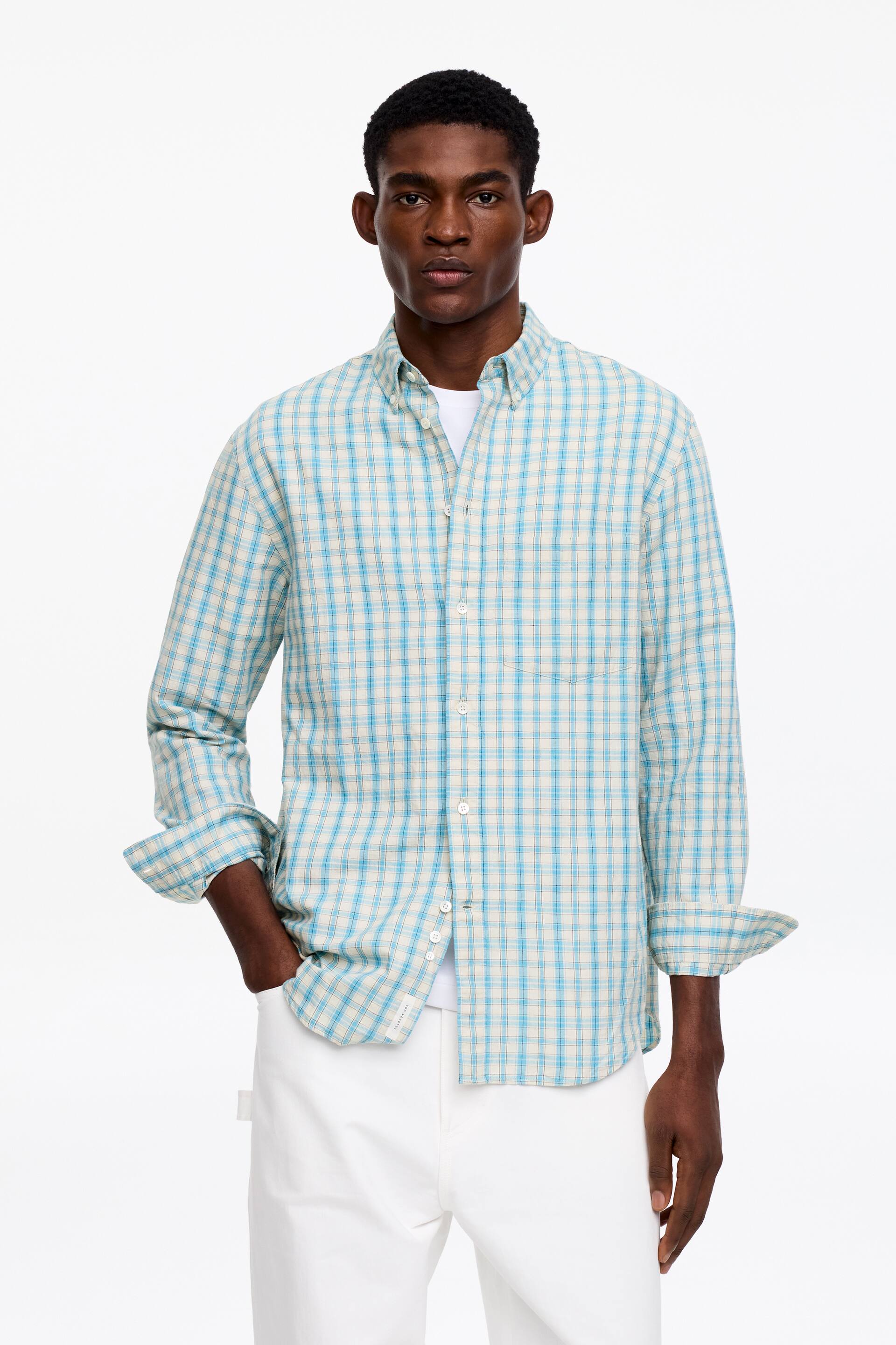 Se større billede: En herreskjorte med knapper har et lyseblåt og off-white ternet mønster, præsenterer en afslappet pasform med en button-down krave og en enkelt brystlomme. Dens lange ærmer er rullet op til underarmene, og den bæres over en hvid t-shirt.