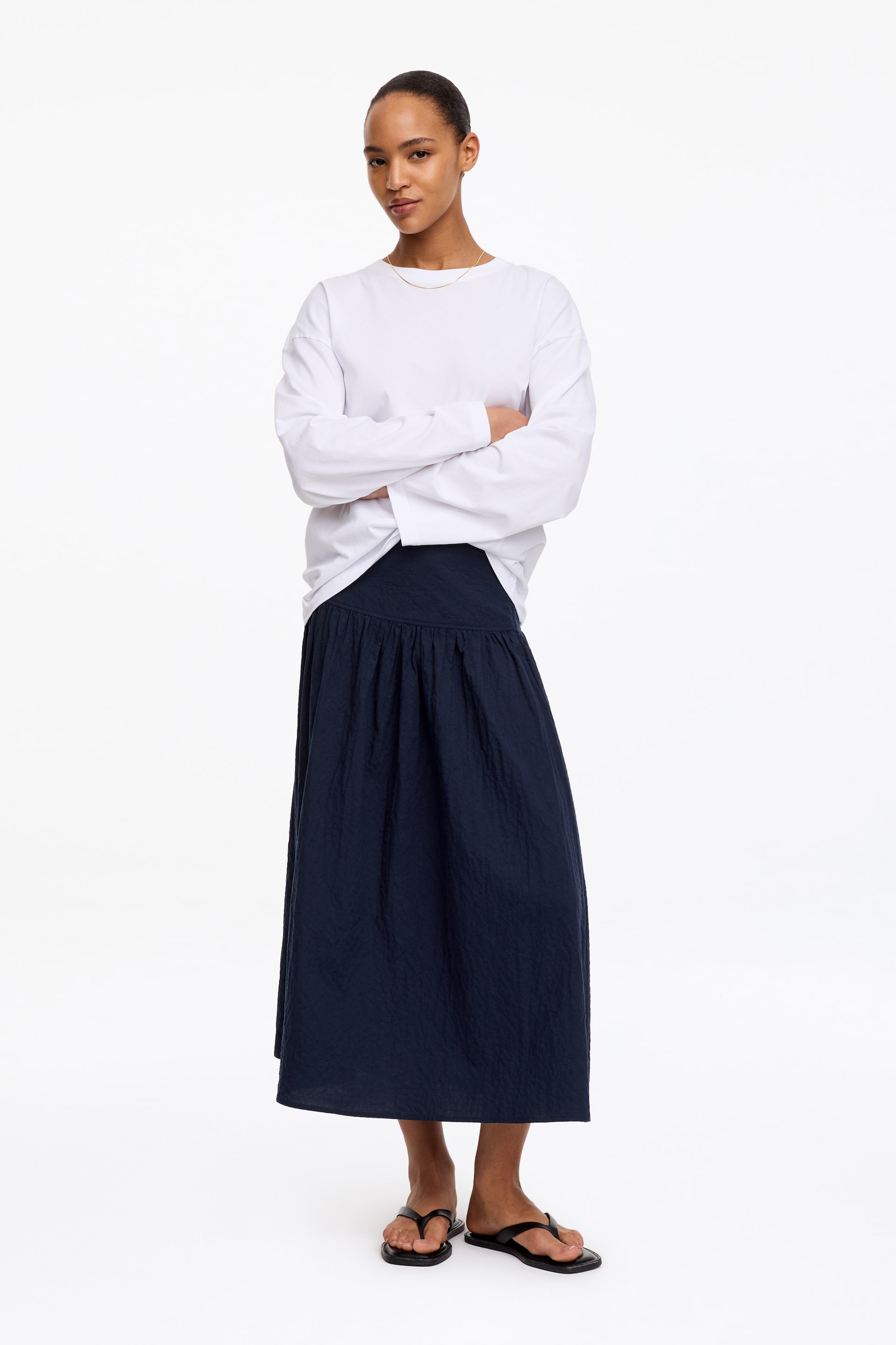 View larger image: A navy midi skirt in cotton seersucker is being worn by the model, displaying a lightly textured, puckered surface and a dropped waist. A white long-sleeved top is being worn tucked into the skirt, creating an elongated silhouette.