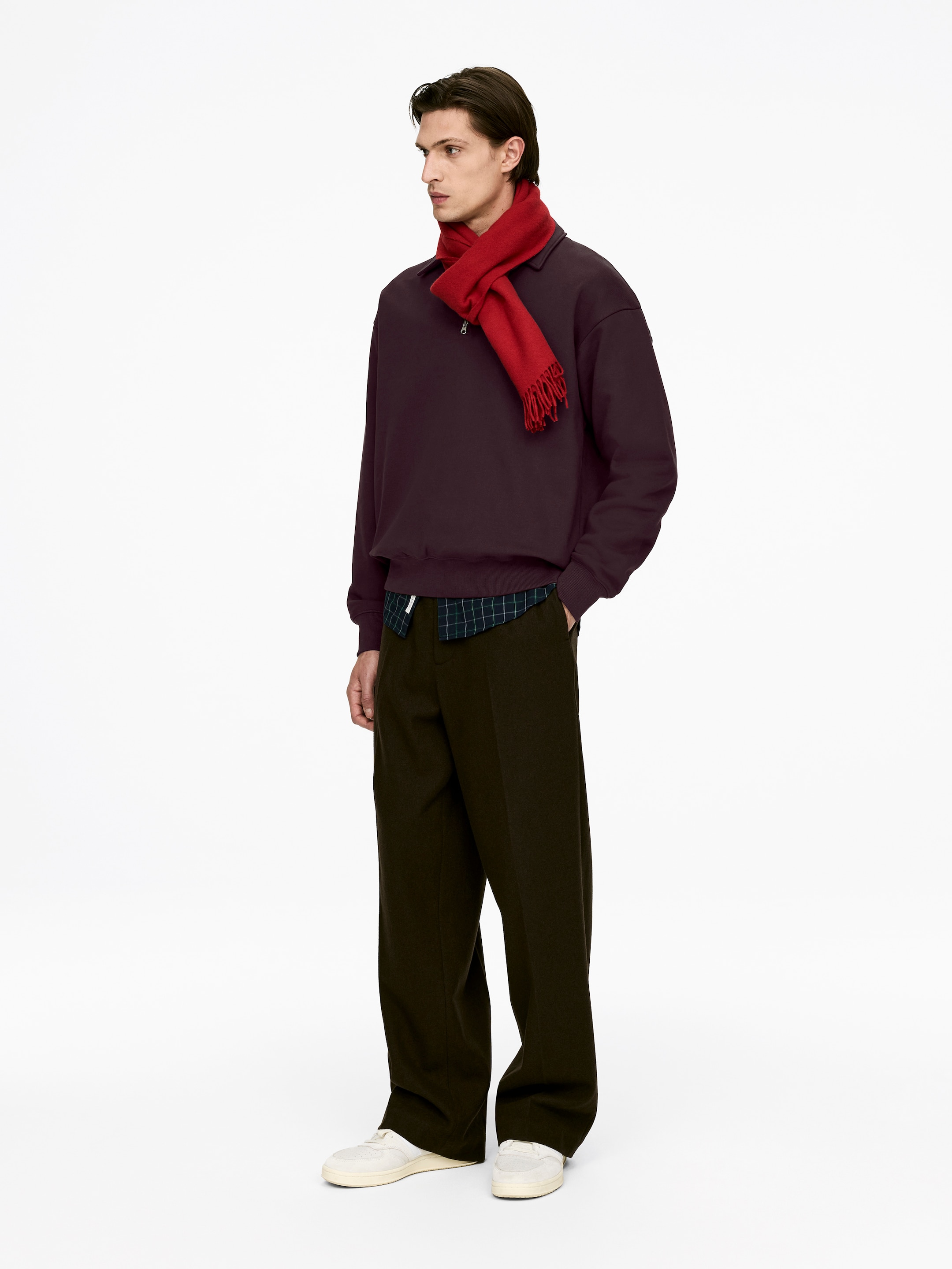 Agrandir l'image: Une tenue présente un polo à fermeture éclair un quart de couleur prune foncé, stylé par-dessus une chemise à carreaux avec son ourlet qui dépasse, et une écharpe rouge à franges drapée autour du cou. L'ensemble est complété par un pantalon large vert olive foncé et des baskets blanc cassé, le tout présenté en vue trois-quarts tourné vers la droite.