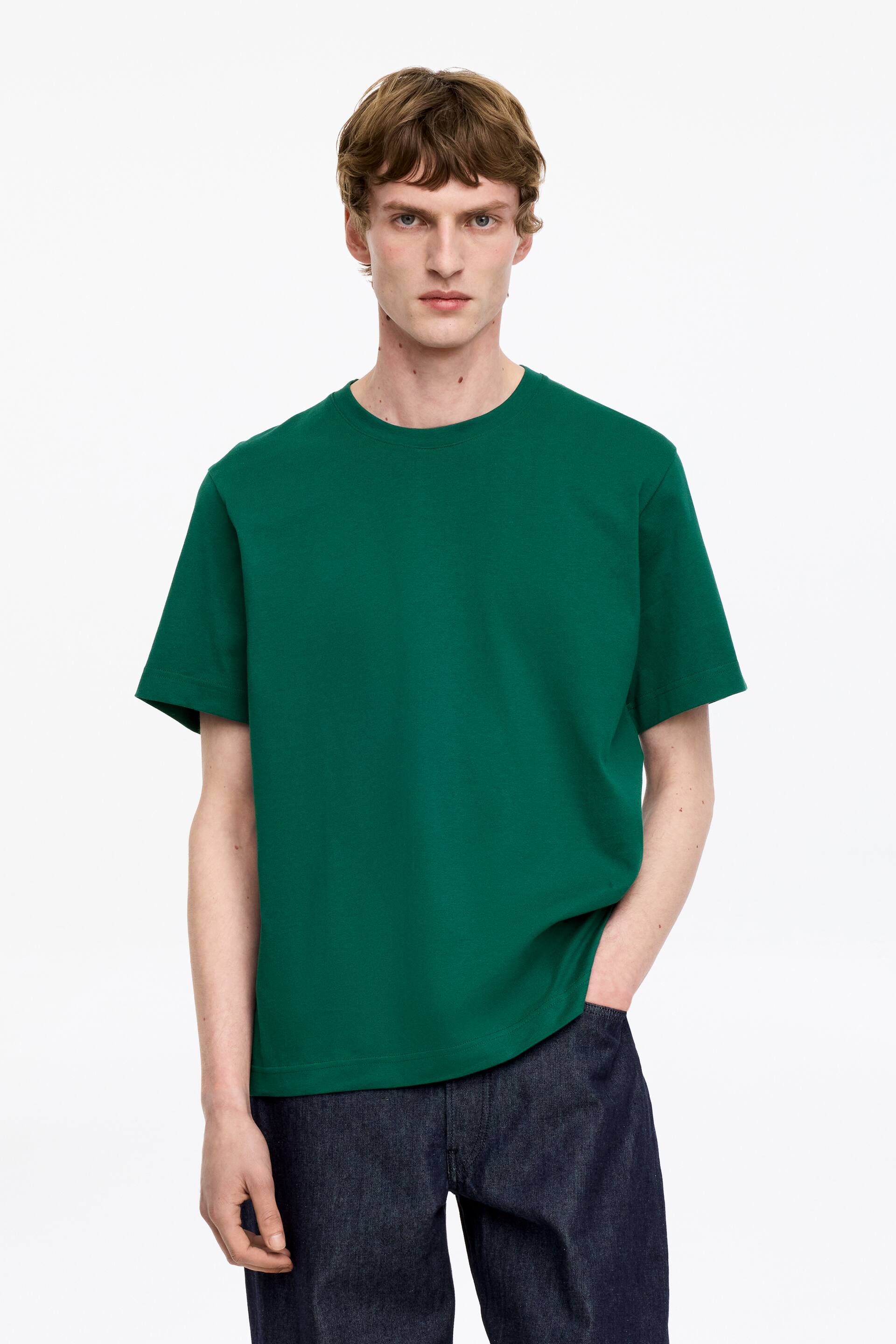 Näytä suurempi kuva: Tummanvihreä t-paita esittelee klassisen pyöreän pääntien ja lyhyet hihat, istuen väljästi vartalolla. Se on yhdistetty tummasävyisiin farkkuihin, ja sen suora helma asettuu lanteille.