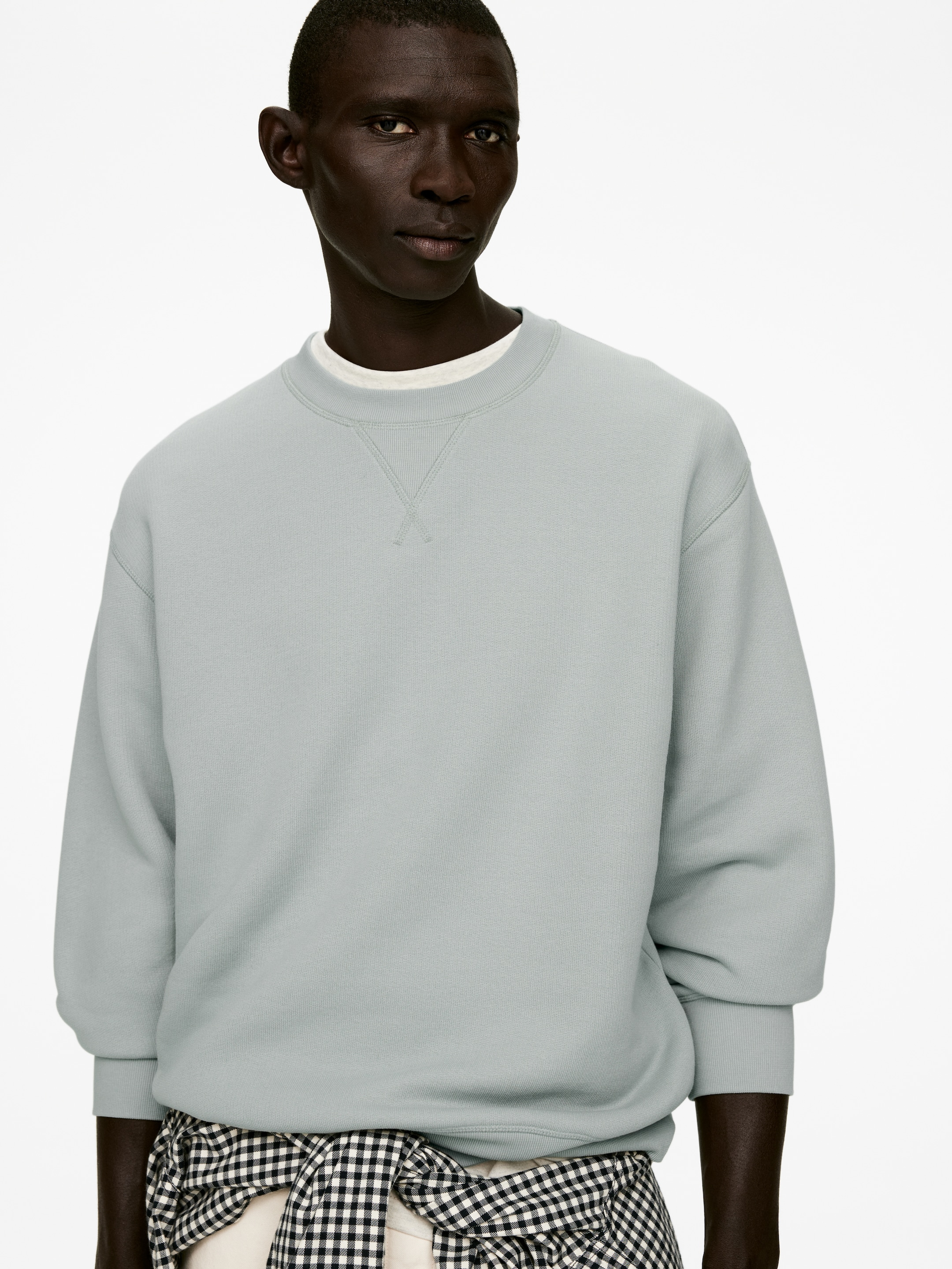 Agrandir l'image: Un sweat-shirt à col rond bleu-gris clair est porté, mettant en valeur sa coupe décontractée vue de face sous un angle légèrement incliné. Il présente un détail en V sous l'encolure, et ses poignets et ourlet côtelés sont visibles, superposés à un t-shirt blanc.
