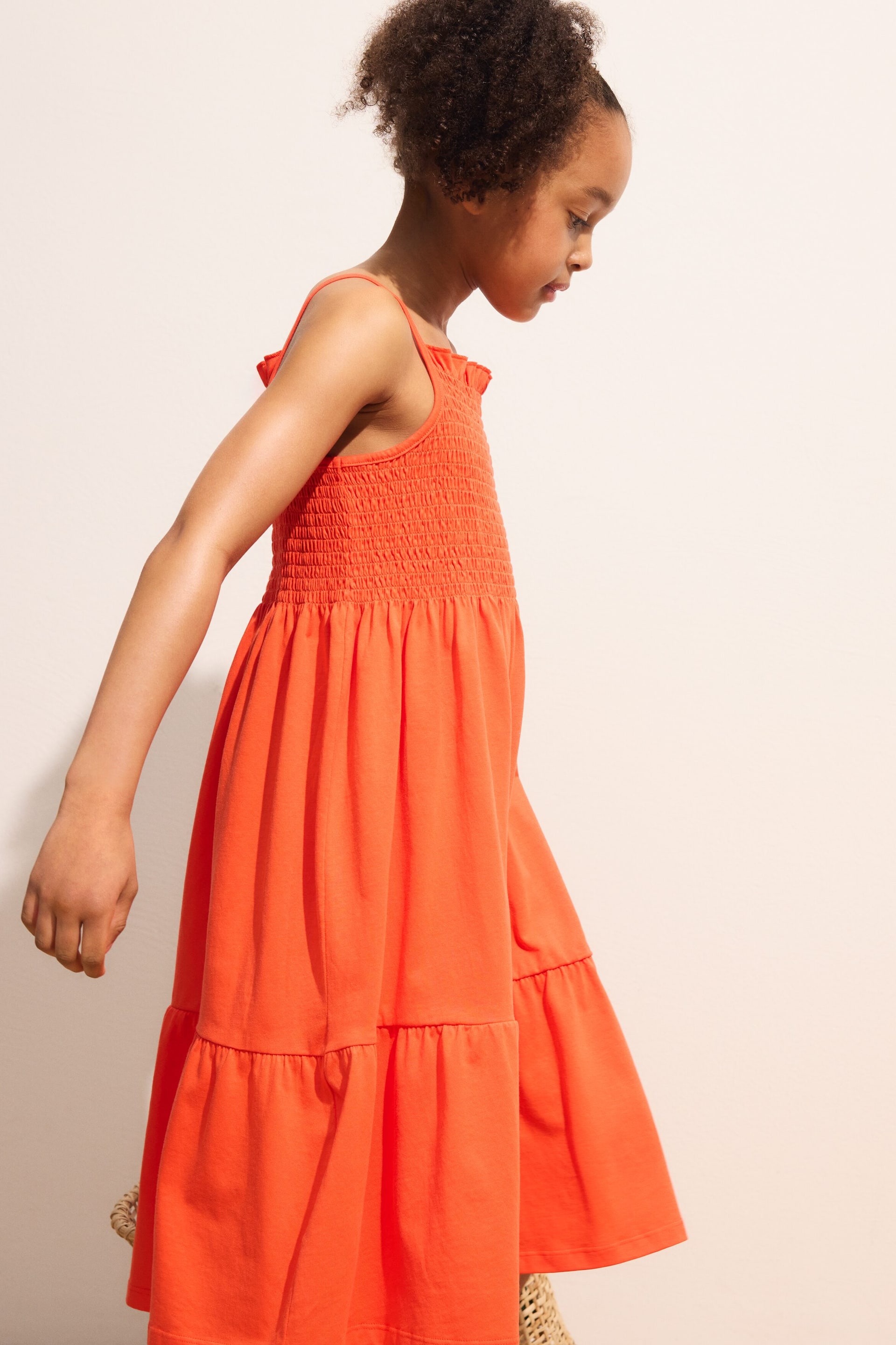 Agrandir l'image: Une robe orange à étages présente un corsage smocké avec de fines bretelles spaghetti et une jupe à deux étages, présentée sous un angle latéral-frontal.