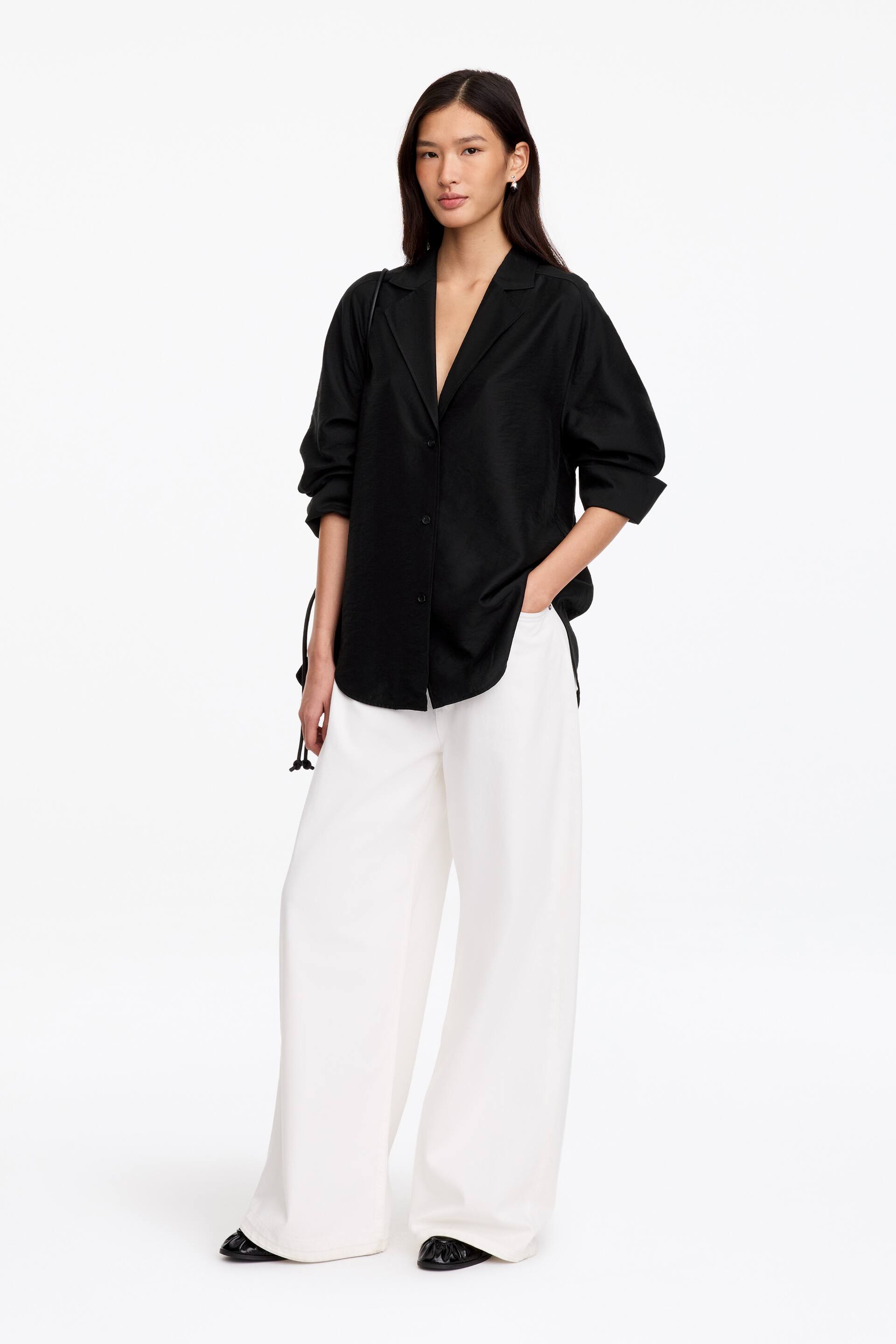 Größeres Bild anzeigen: Ein schwarzer, ungefütterter Blazer mit fallendem Revers, Knopfleiste und abgerundetem Saum wird vom Model getragen. Der Blazer wird mit einer weißen Wide-Leg-Hose kombiniert, wobei das Model eine Hand lässig in die Hosentasche gelegt hat.