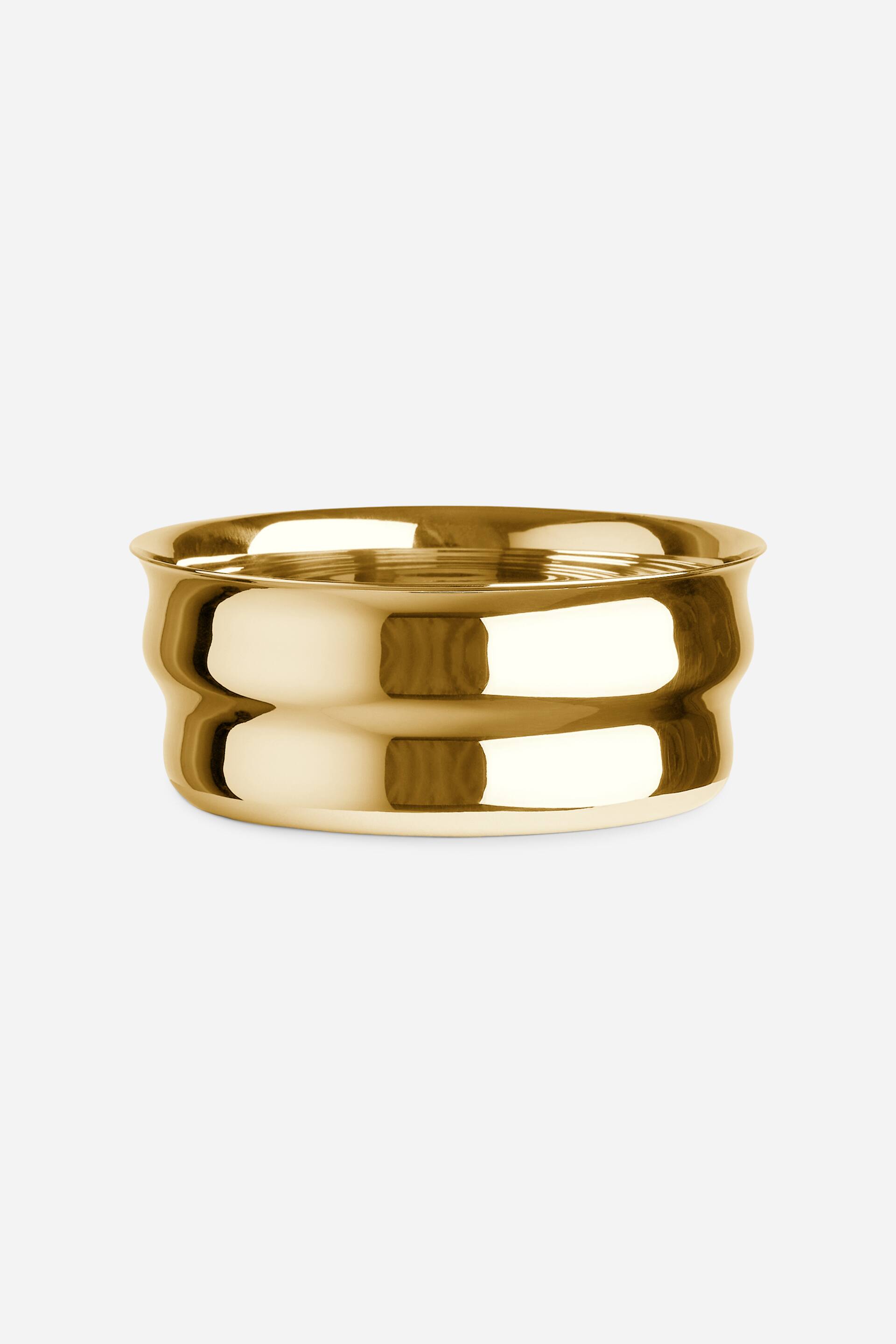 View larger image: A brass bottle coaster is displayed from a front-facing angle, featuring a highly polished, reflective gold-toned finish. The coaster has a cylindrical design with a distinctive double-contoured, undulating side profile and a slightly flared top rim.