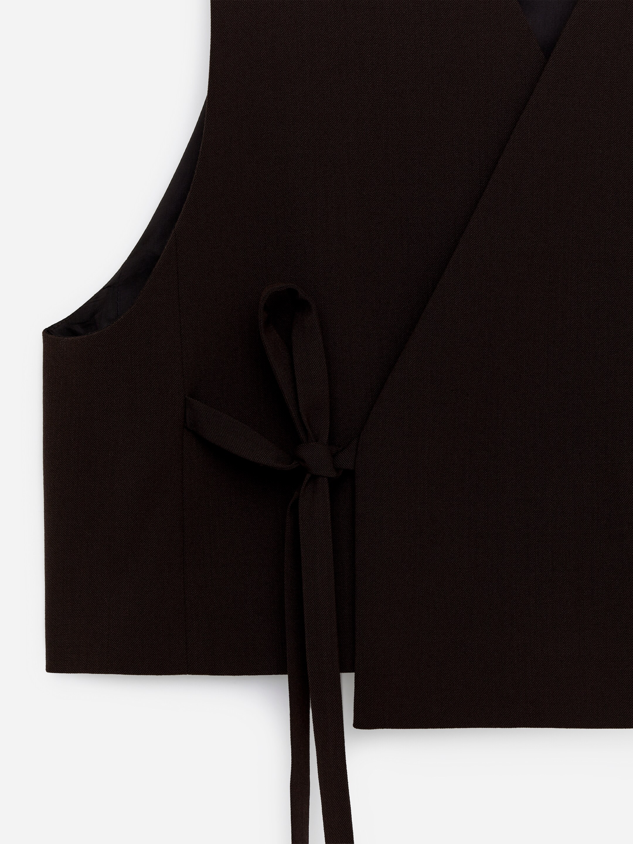 Agrandir l'image: Un gilet ajusté de couleur sombre présente son DEVANT, avec une fermeture superposée. Une sangle de cravate sécurise le devant, nouée sur le côté gauche, et un rabat de poche décoratif est visible sous la cravate.